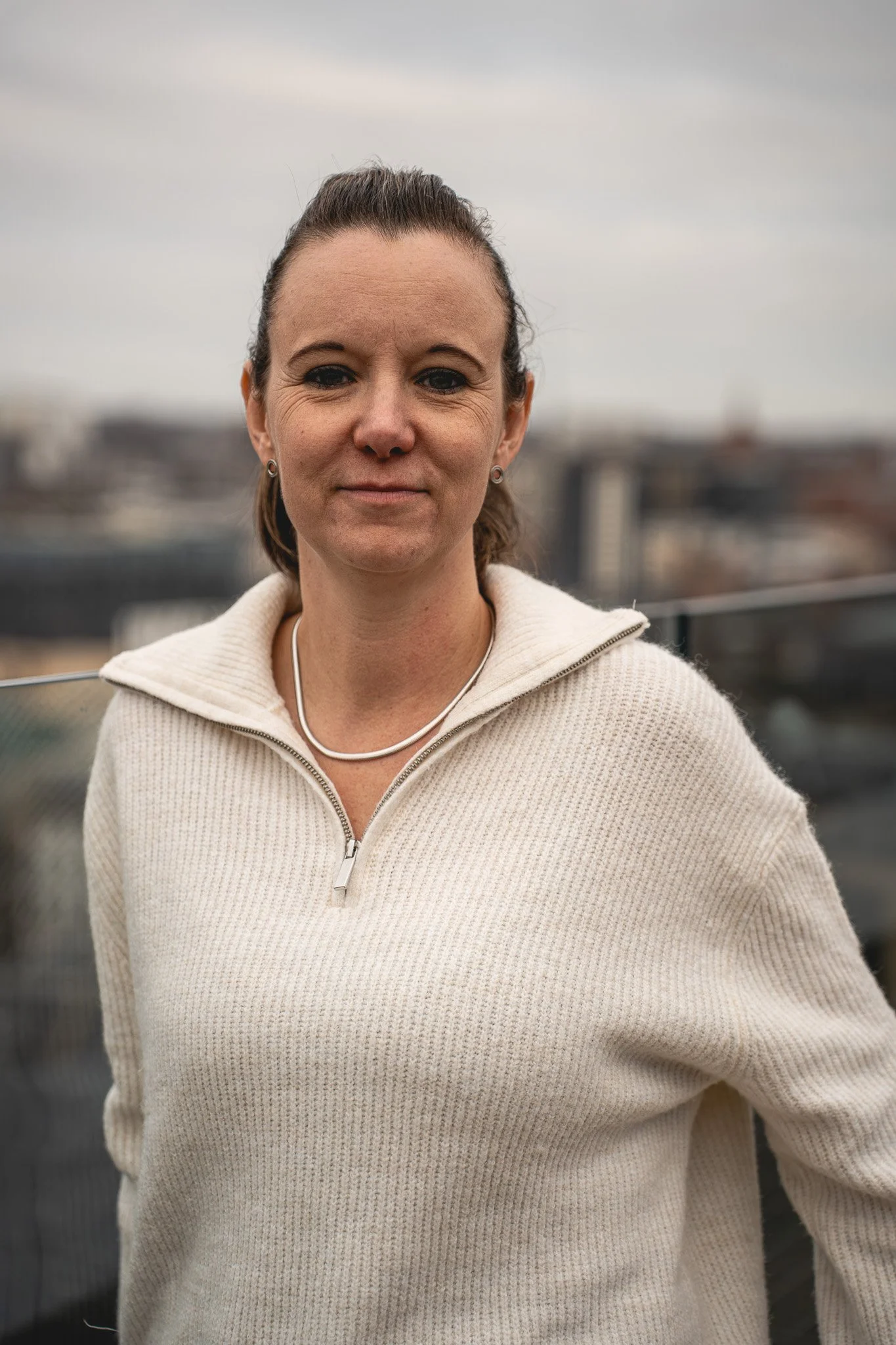 Charlie Bartle, COO and Co-Founder of Zygens with brown hair tied back, wearing a cream knit sweater with a zipper, standing outdoors on a cloudy day with a Leeds cityscape blurred in the background.