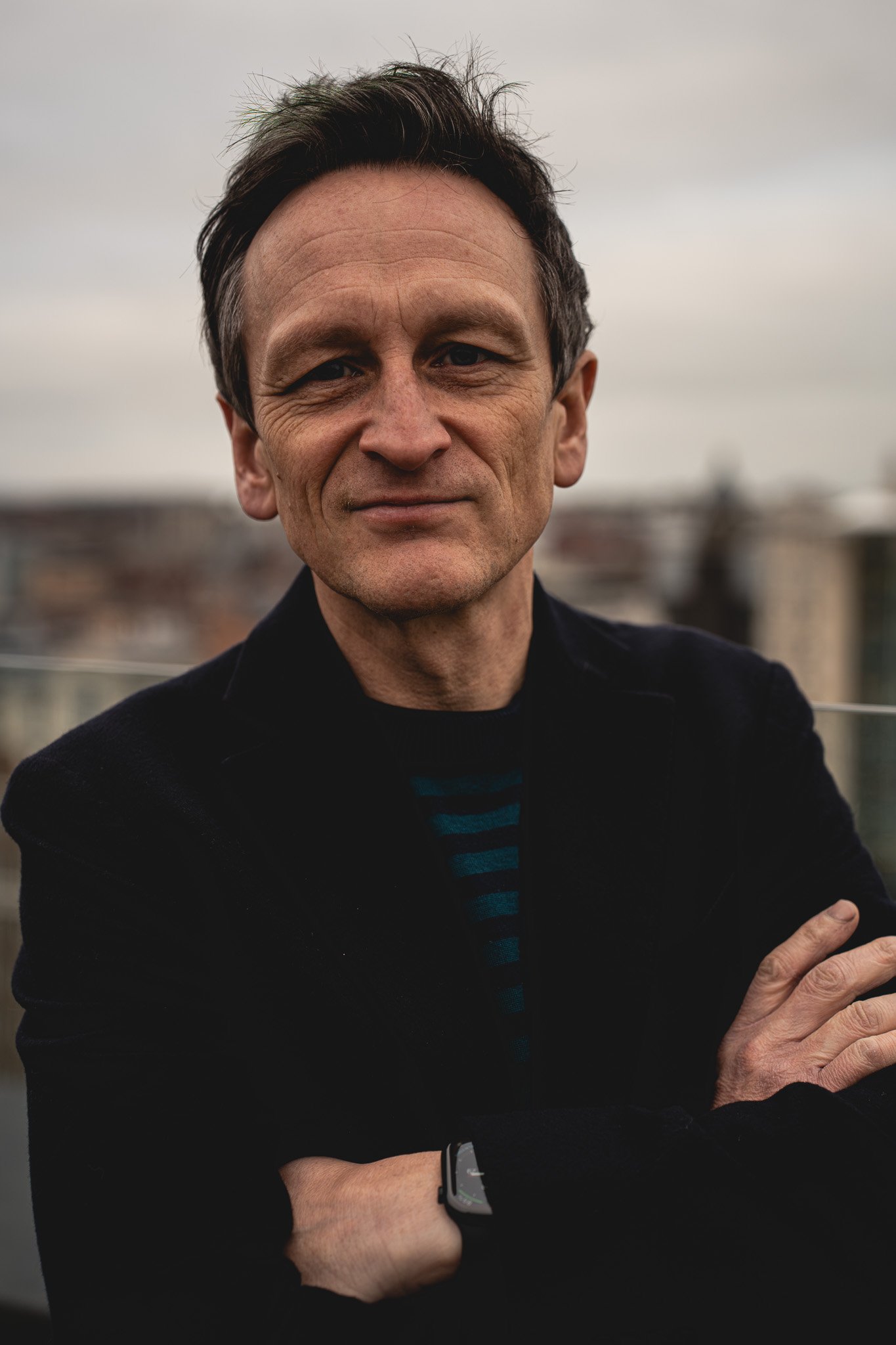 Andy Roberts, CMO and Co-Founder of Zygens, shown with dark hair and light skin, wearing a navy blue jacket and striped jumper, standing outdoors with a Leeds cityscape in the background.
