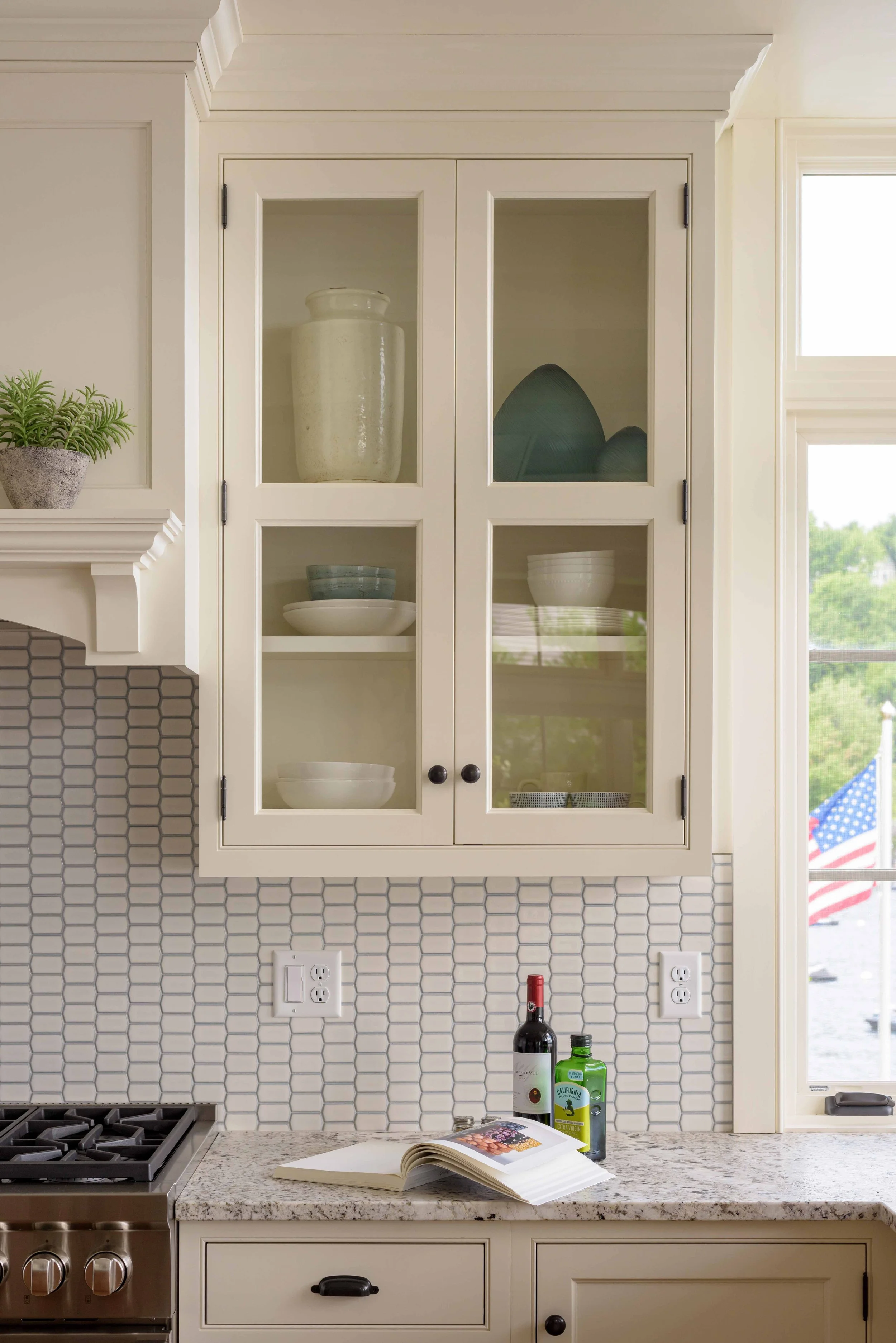 Kitchen cabinet with glass panes, open to reveal bowls and decorative plates, over a countertop with an open cookbook, wine bottle, and olive oil bottle, next to a window with an American flag outside.