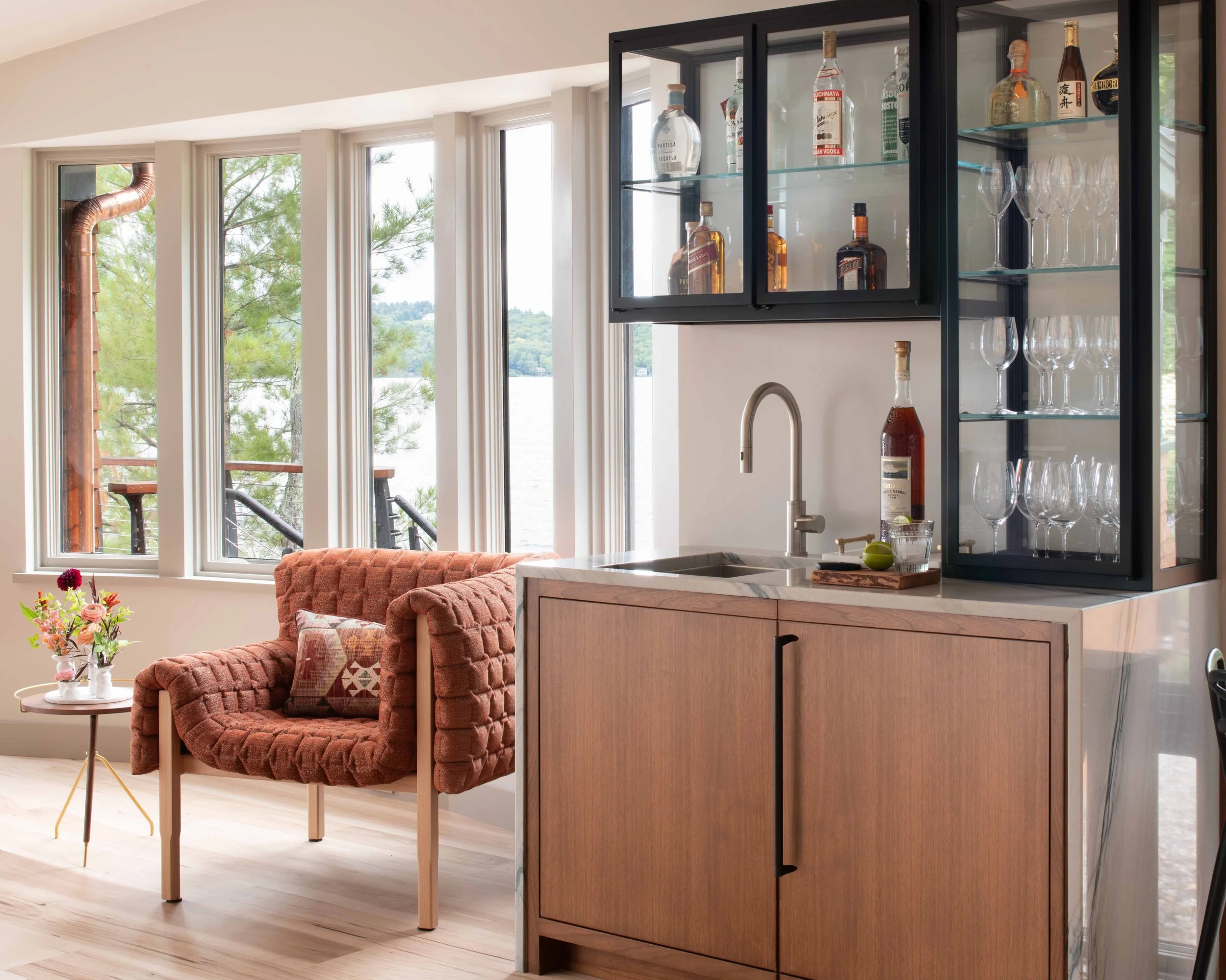 A cozy living space with large windows overlooking a lake, a plush reddish-brown armchair with a patterned pillow, a small glass side table with a vase of flowers, and a kitchen area featuring a black cabinet with glass doors displaying liquor bottles and glassware, wood cabinets, and a stainless steel faucet.