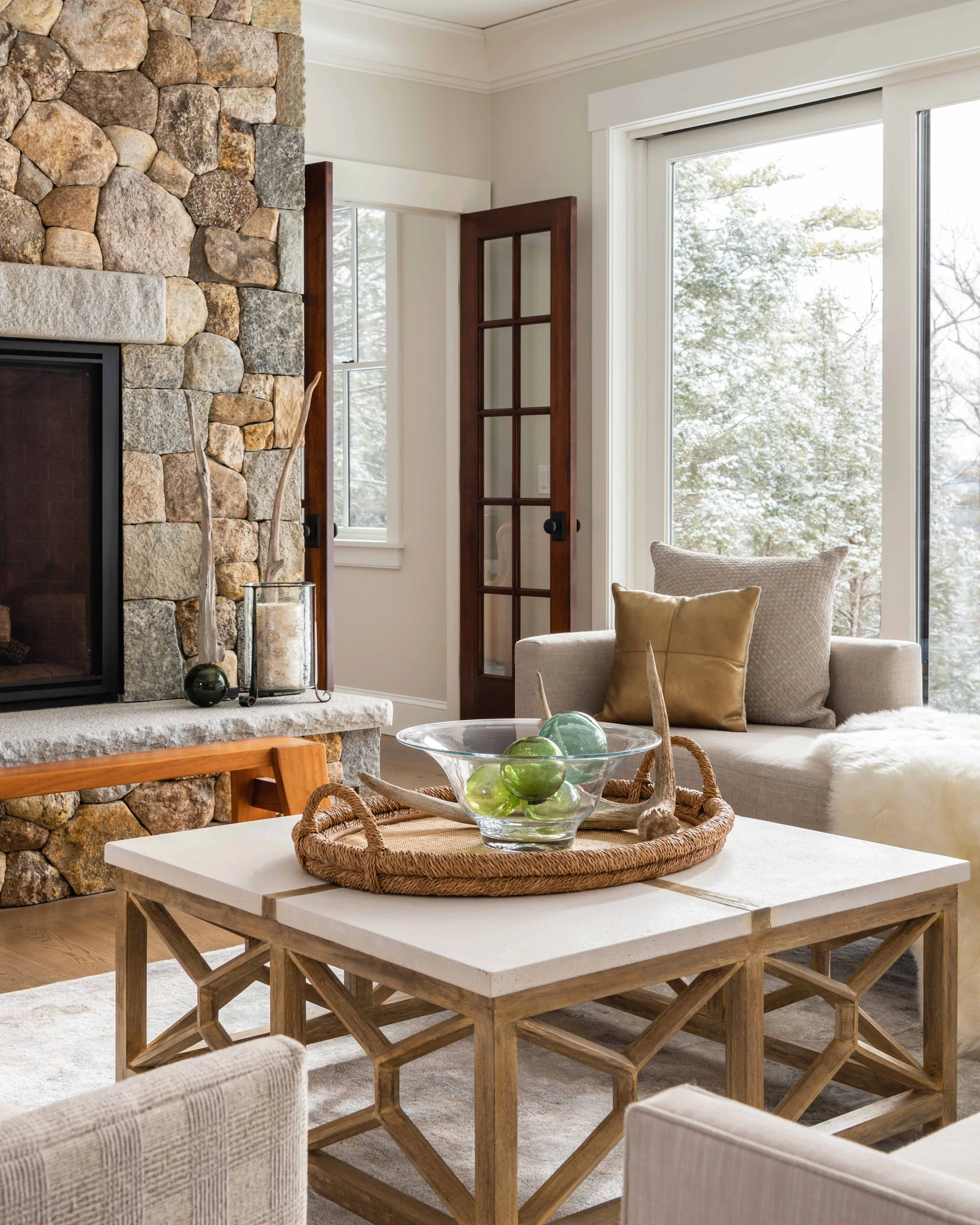 Living room with stone fireplace, wooden-framed glass door, beige armchair, decorative cushions, glass bowl with green and purple decorative balls, and large window showing snow-covered trees outside.