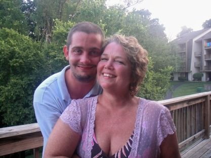 A young man and an older woman smiling and hugging on a wooden deck, with greenery and a building in the background.