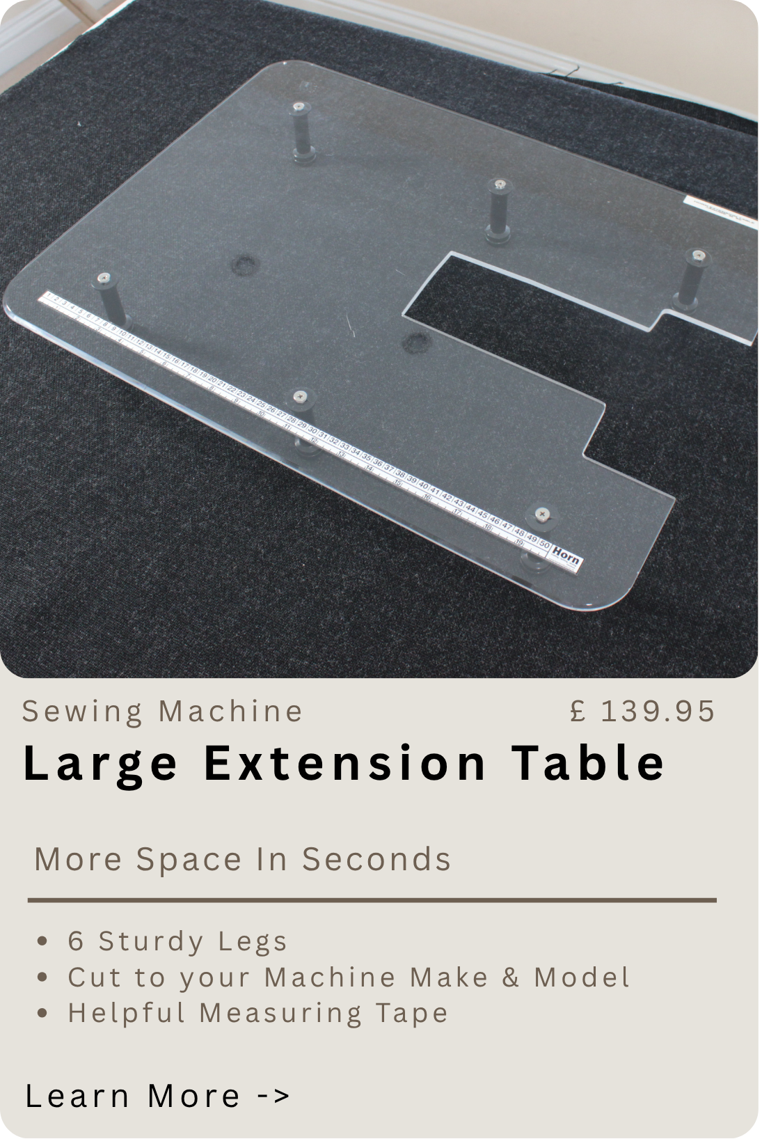 A large, clear plastic extension table with four black legs attached, placed on a black carpeted surface. The table has measurement markings along the edge and is designed to extend the work surface of a sewing machine.