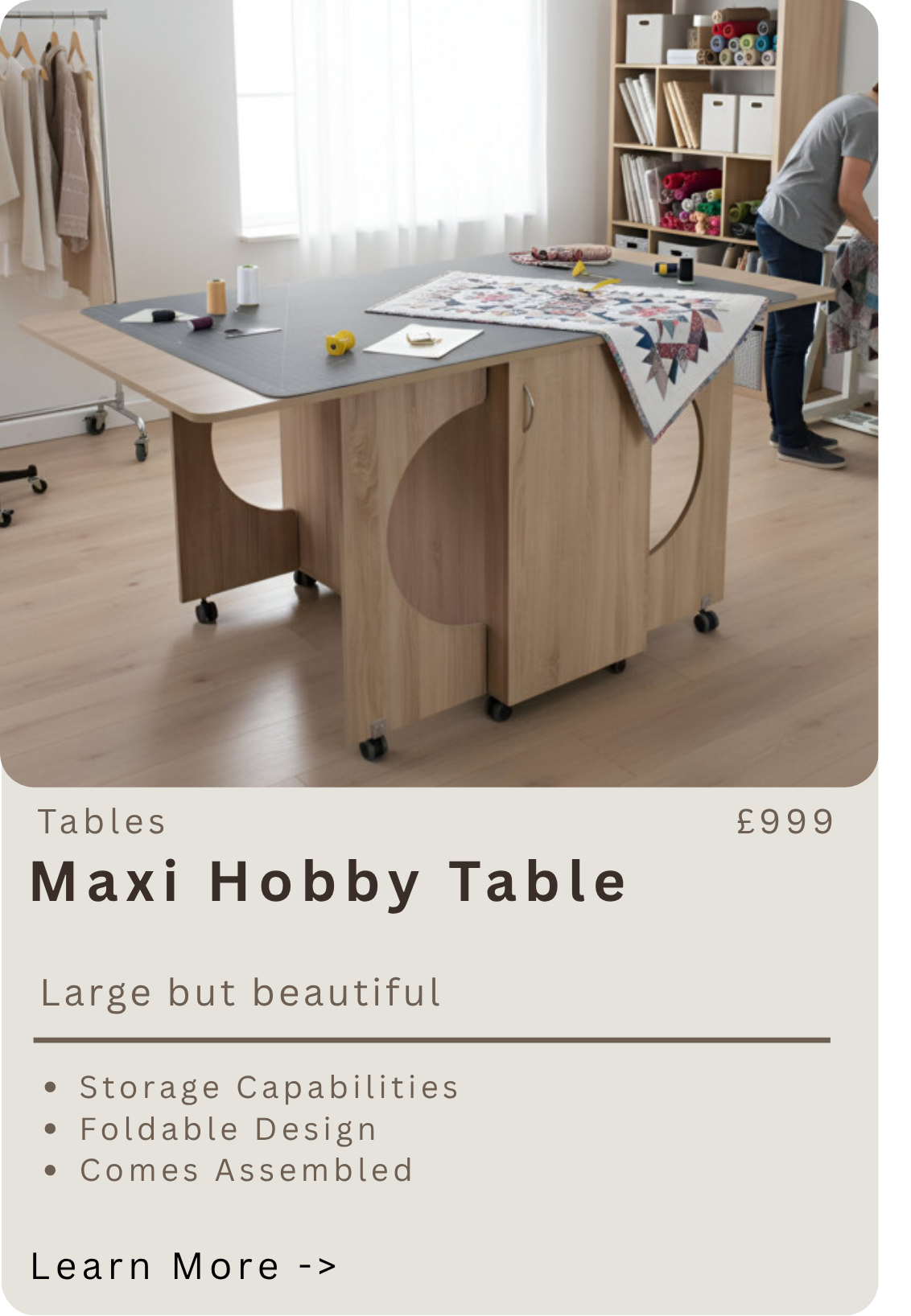 A large wooden hobby table with storage shelves in a bright room. The table holds fabric, sewing tools, and scissors, with a person working on a sewing project nearby.