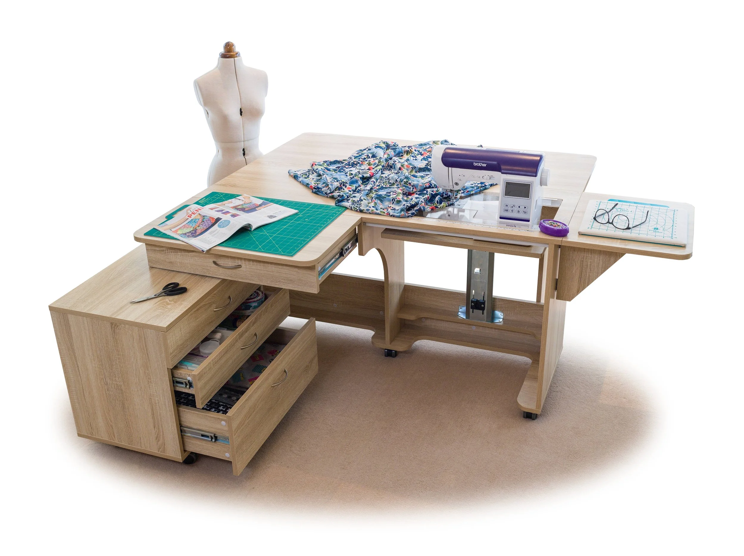 A sewing workspace with a wooden desk, the desk has an open drawer with sewing supplies. There is a mannequin torso in the background, a sewing machine, fabric, scissors, and a thread spool on the desk. An open magazine, a cutting mat, and a pair of