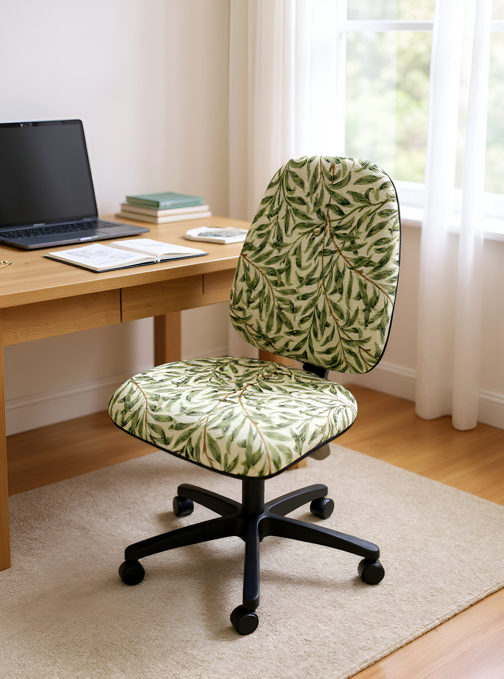 An office chair with a leafy green plant pattern, placed on a beige rug in front of a wooden desk with a laptop, notebooks, and a phone. The background has a window with sheer white curtains and outdoor greenery.