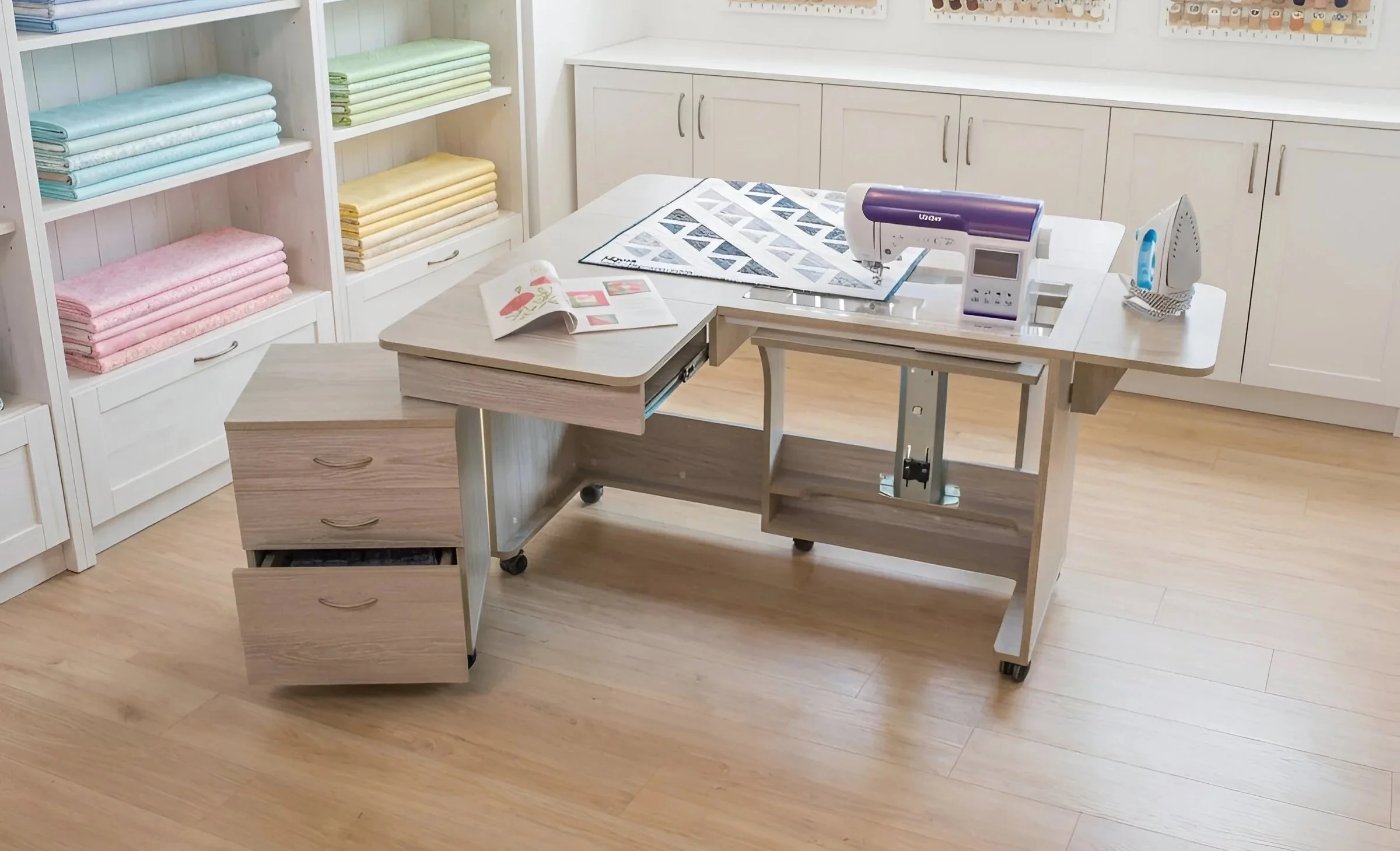 A crafting room with a white table, sewing machine, iron, and open book. Shelves with pastel-colored folded fabrics are in the background.