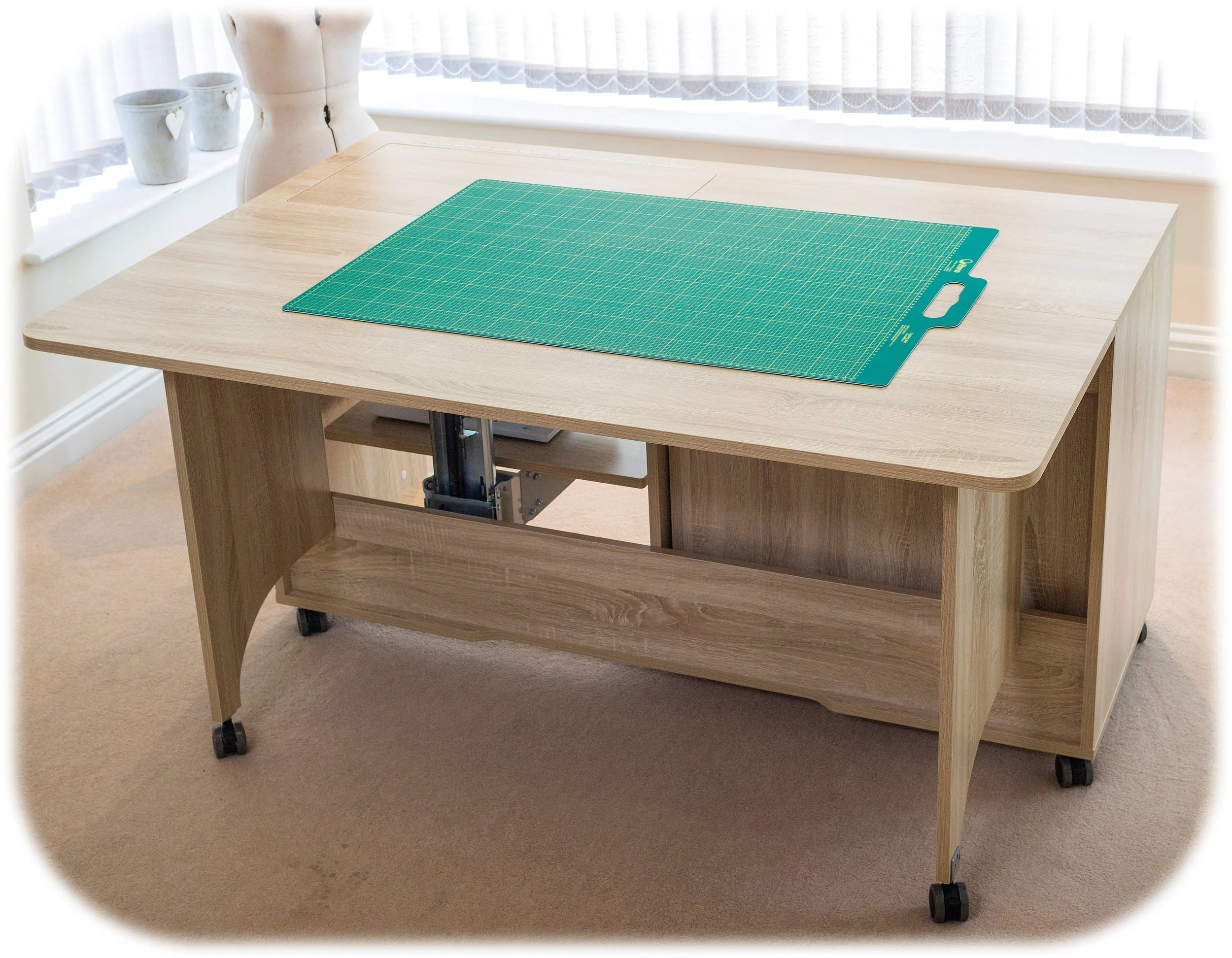 A wooden drafting table with adjustable height, on wheels, placed in a room with beige carpet and curtains. A green cutting mat is on top.
