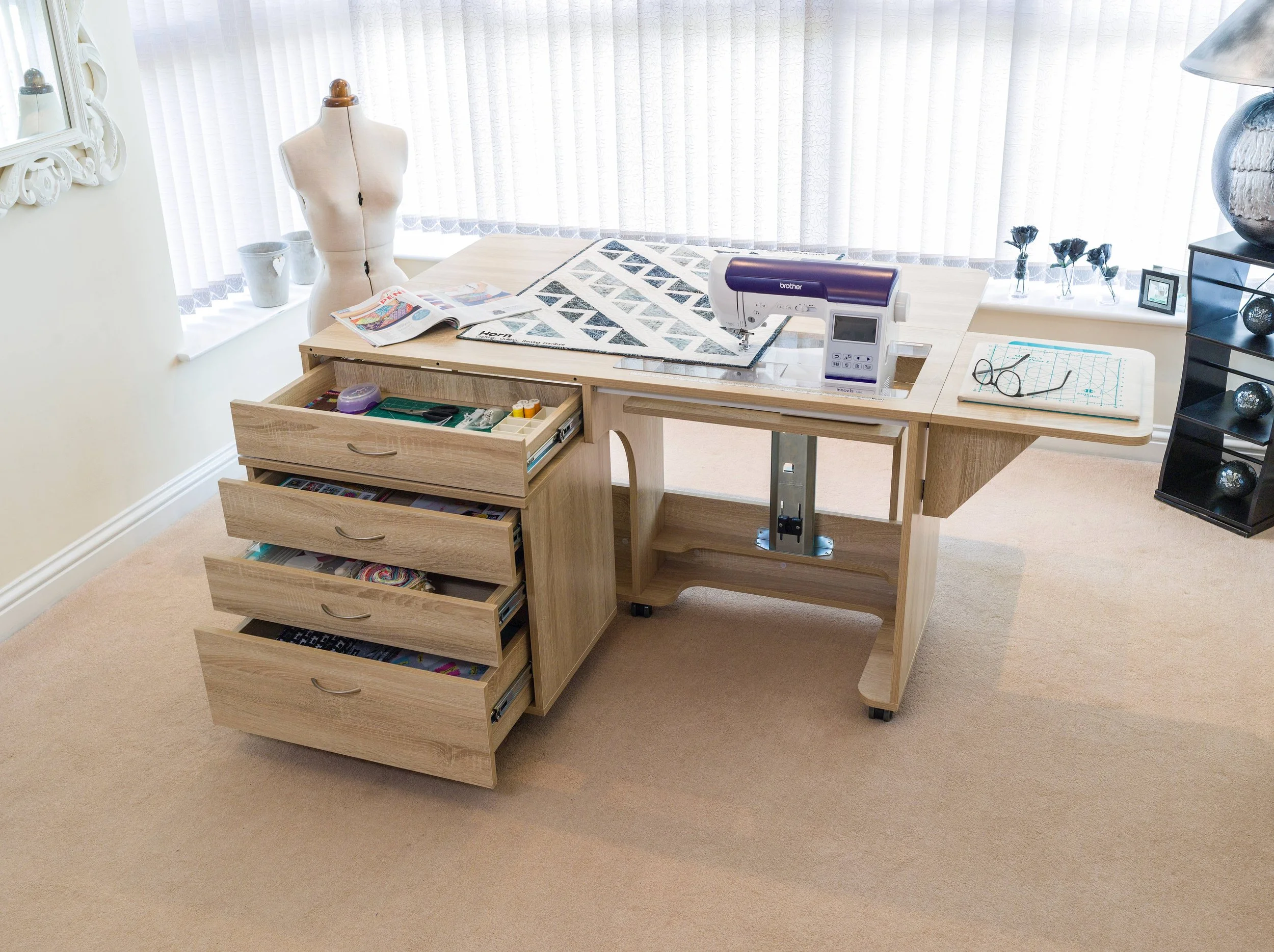 A sewing workspace with a wooden table, a mannequin, and drawers filled with sewing supplies. There's a sewing machine on the table, open magazines, and a grid pattern with glasses. The room has large windows with sheer curtains and decorative items