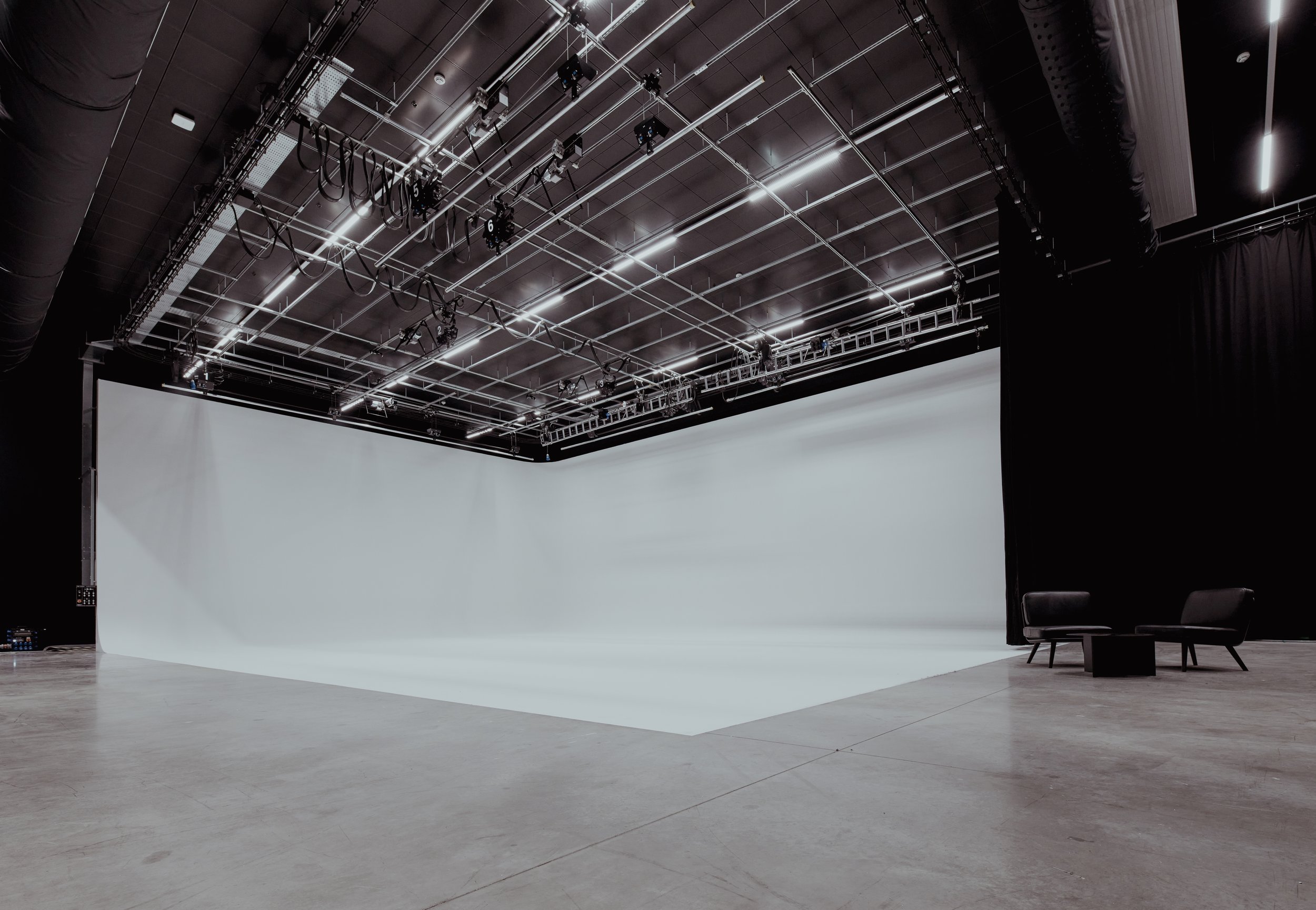 A large empty studio with a white cyc wall, black curtains on the side, and two black chairs with a small black table between them.