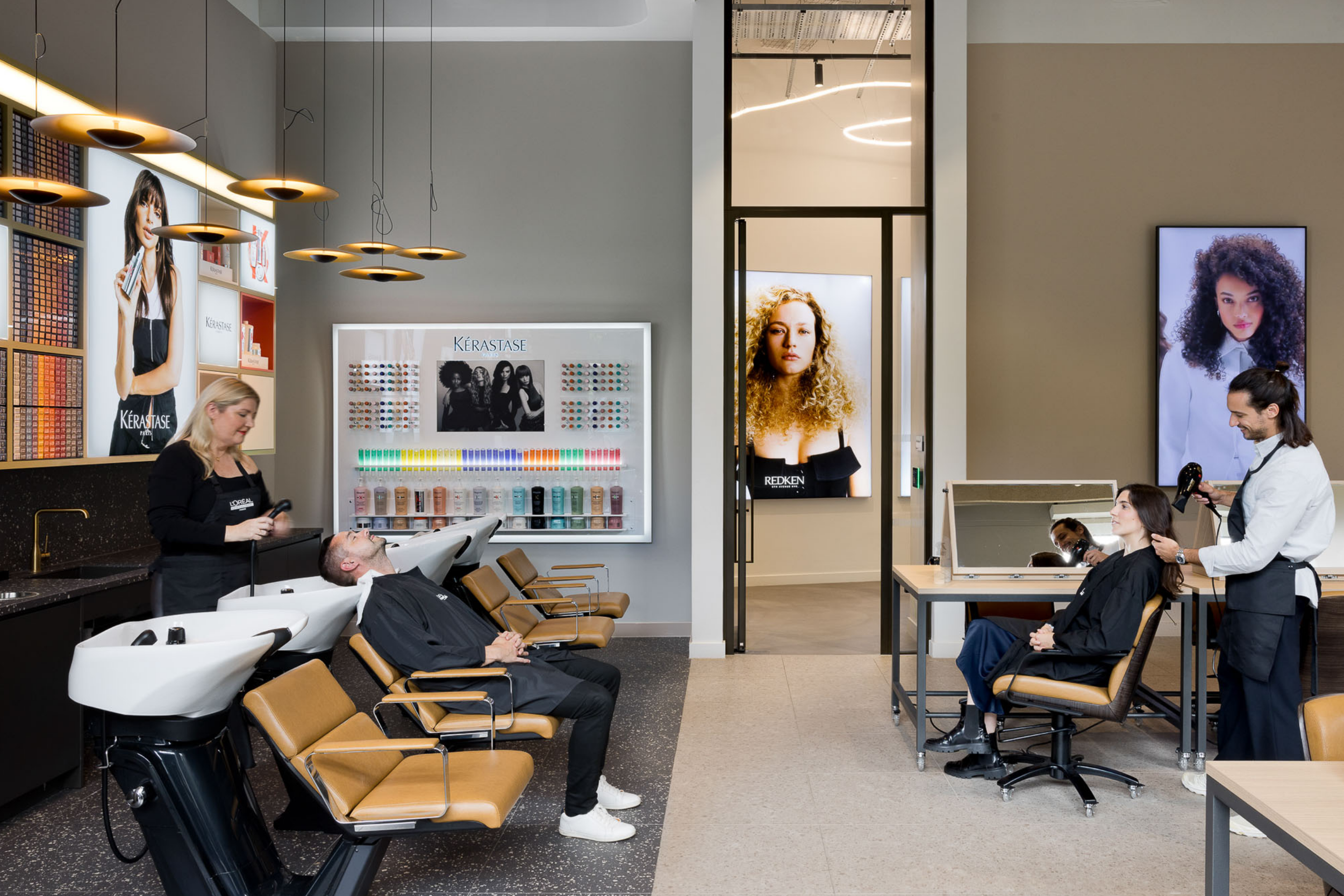 A modern hair salon with a man getting a haircut by a stylist, a customer receiving a hair wash, and large digital screens displaying images of women with different hairstyles.