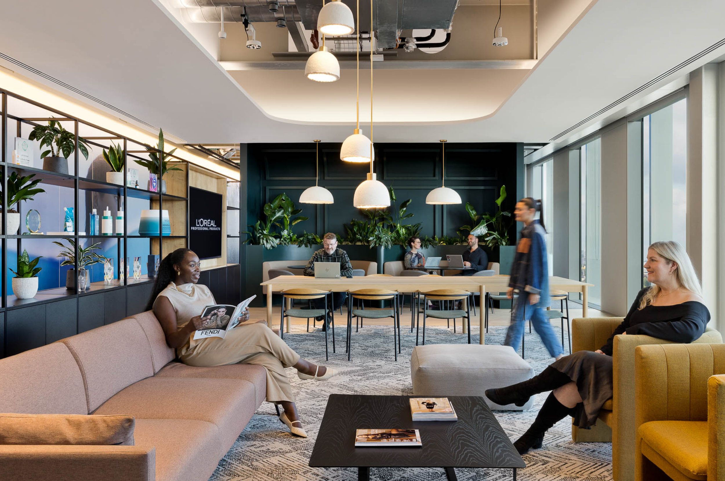 Modern office lounge with people sitting and working on laptops and reading magazines, decorated with plants, shelves, and large windows.