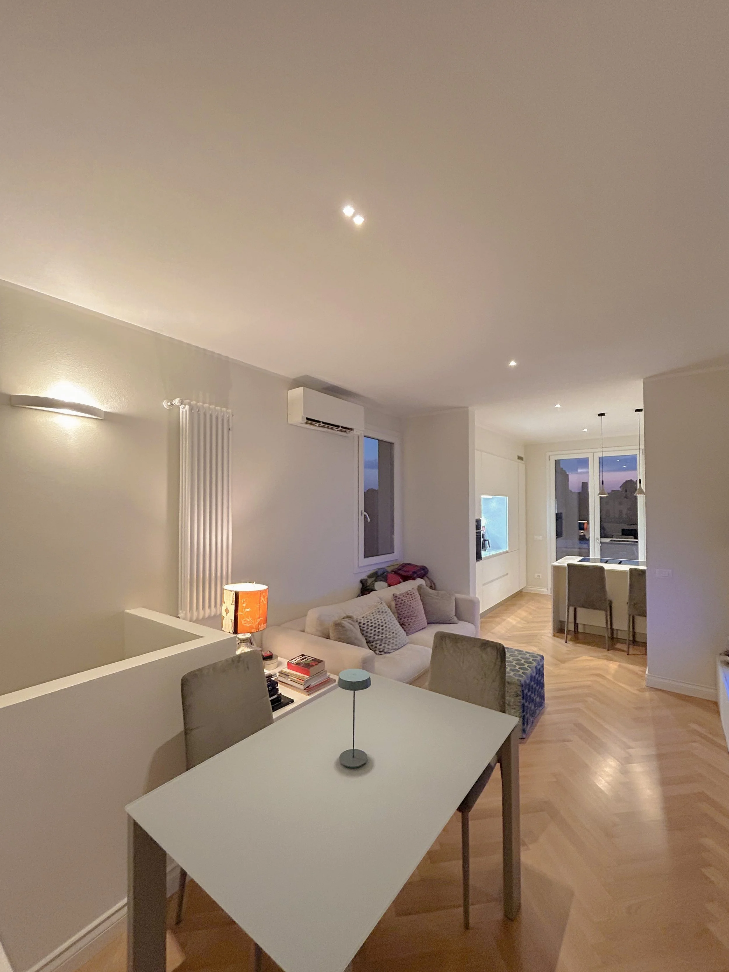 Living room with white walls, hardwood floors, a white couch with pillows, a table with books and a lamp, and large windows with evening sky visible.
