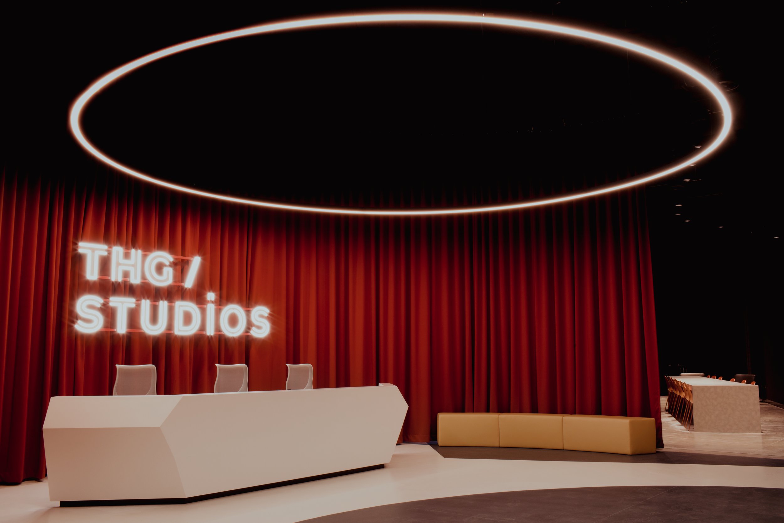 A modern conference room with a white angular desk and four chairs behind it, red curtains, a neon sign reading 'THG STUDIOS,' a circular light fixture on the ceiling, and a beige curved seating area to the right.
