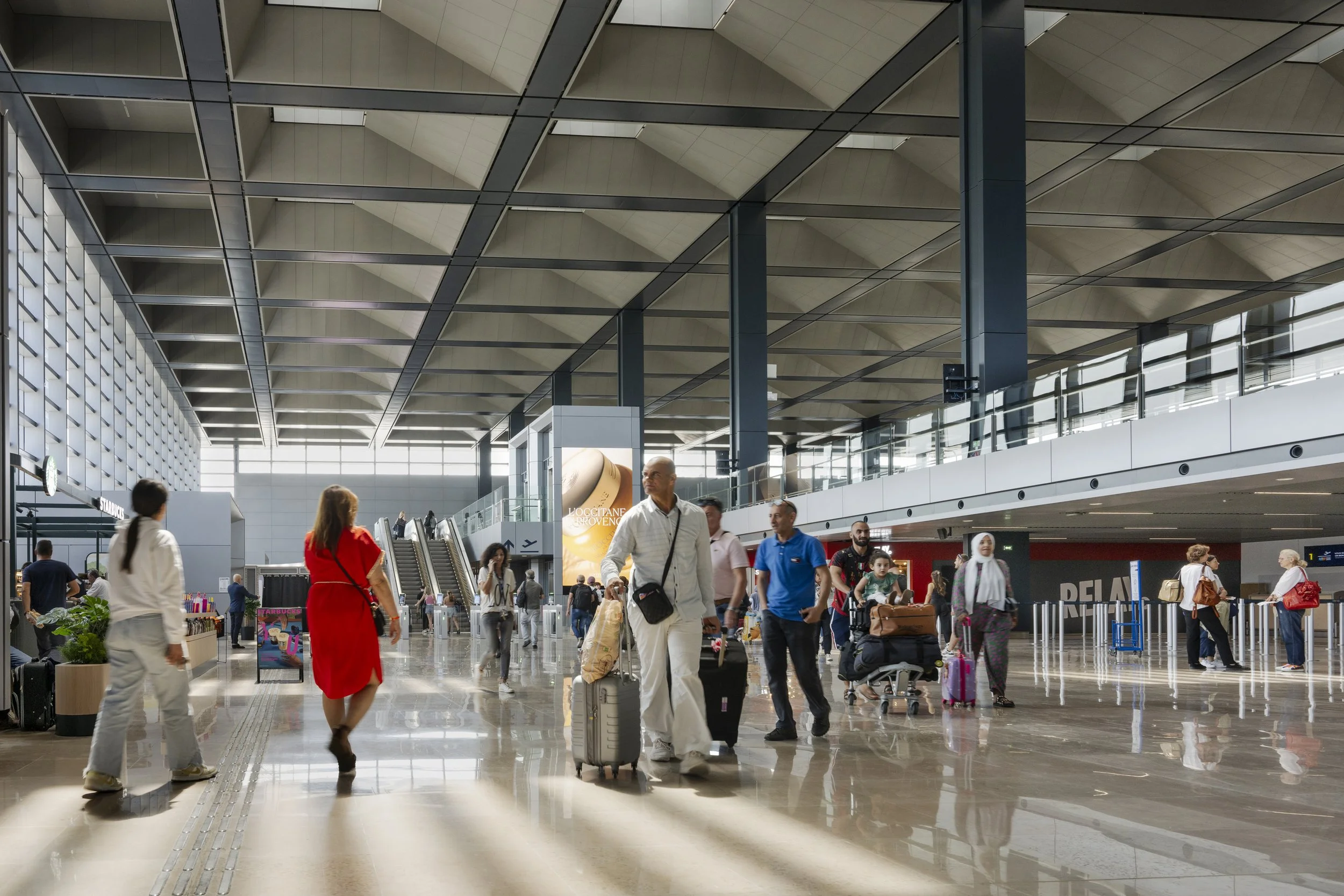 People walking through an airport terminal with luggage and carts, high ceiling with modern architecture, bright natural light from large windows.
