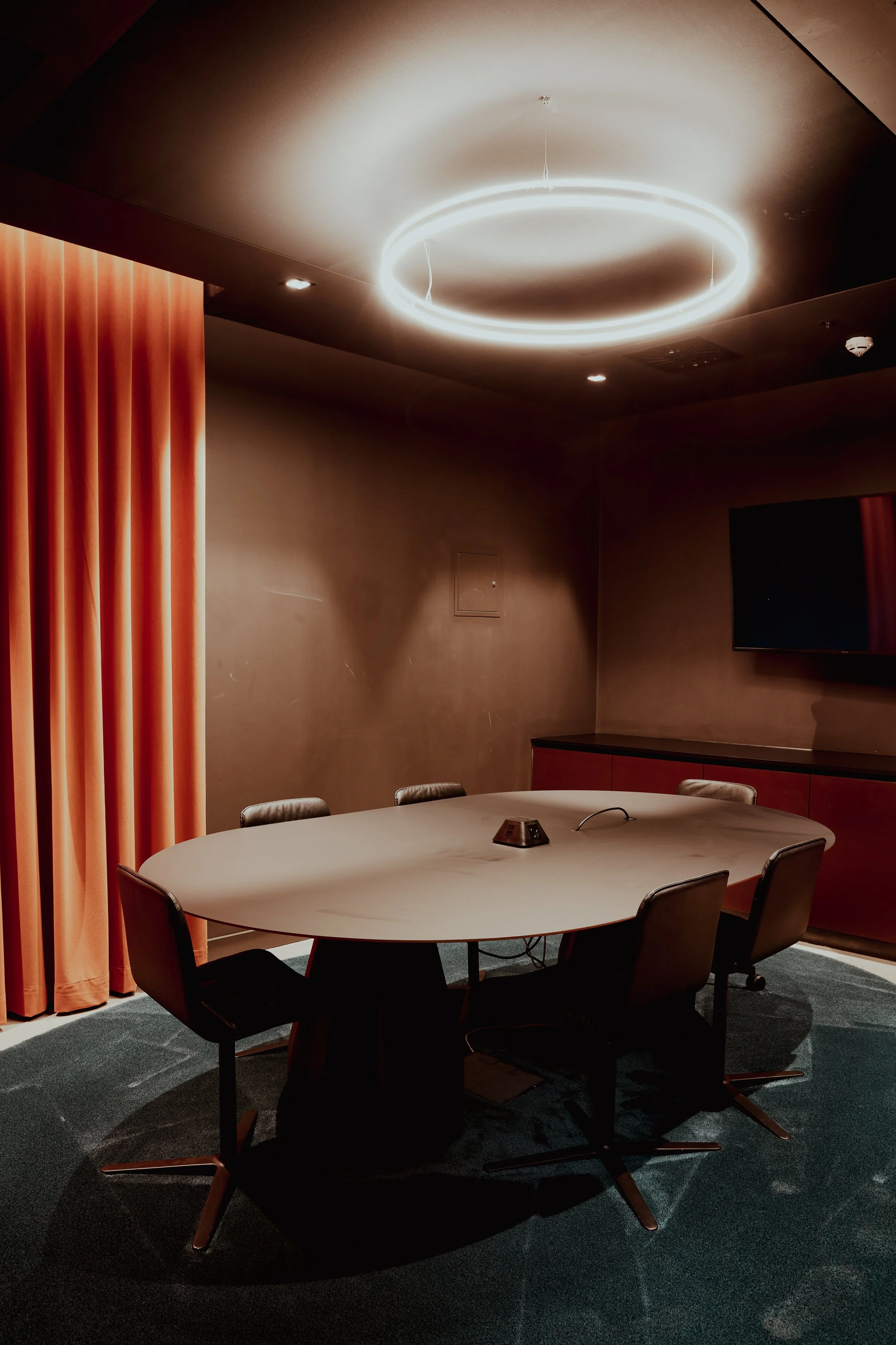 Empty conference room with a round table, six chairs, a large TV on the wall, and a modern circular ceiling light.