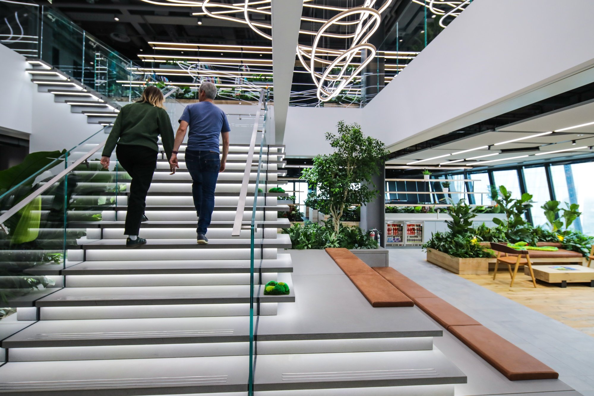 Two people walking up illuminated modern staircase inside a building with plants and large windows.