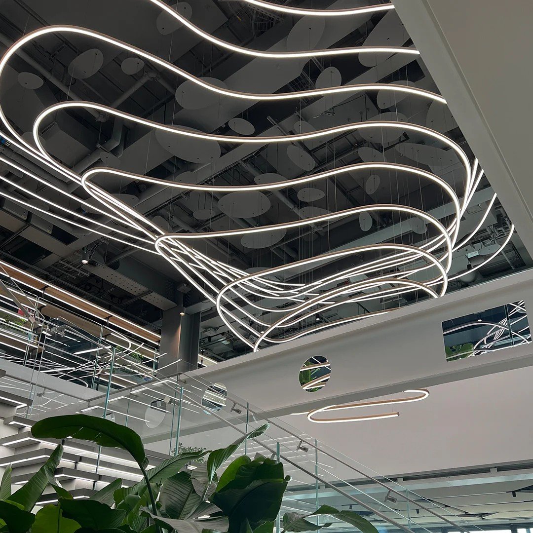 Modern interior with abstract white LED light fixtures hanging from a high ceiling, glass railings, and large green plants in the foreground.