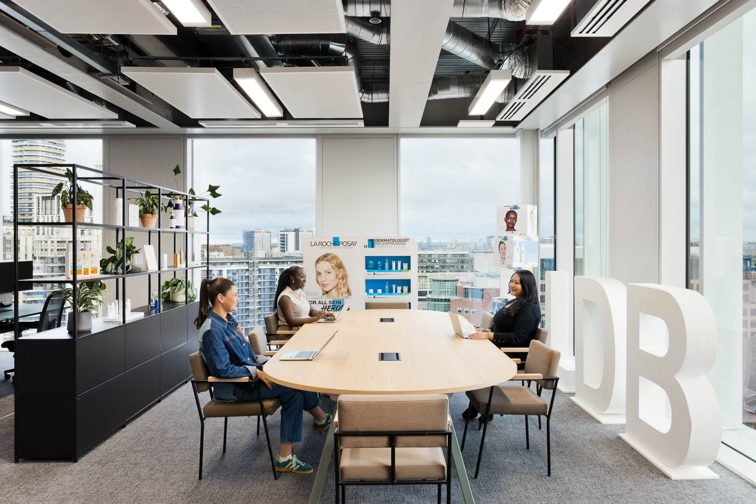 People in a conference room with large windows and city skyline, three women seated at a long table, two women working on laptops, informational posters for skincare products, large white 