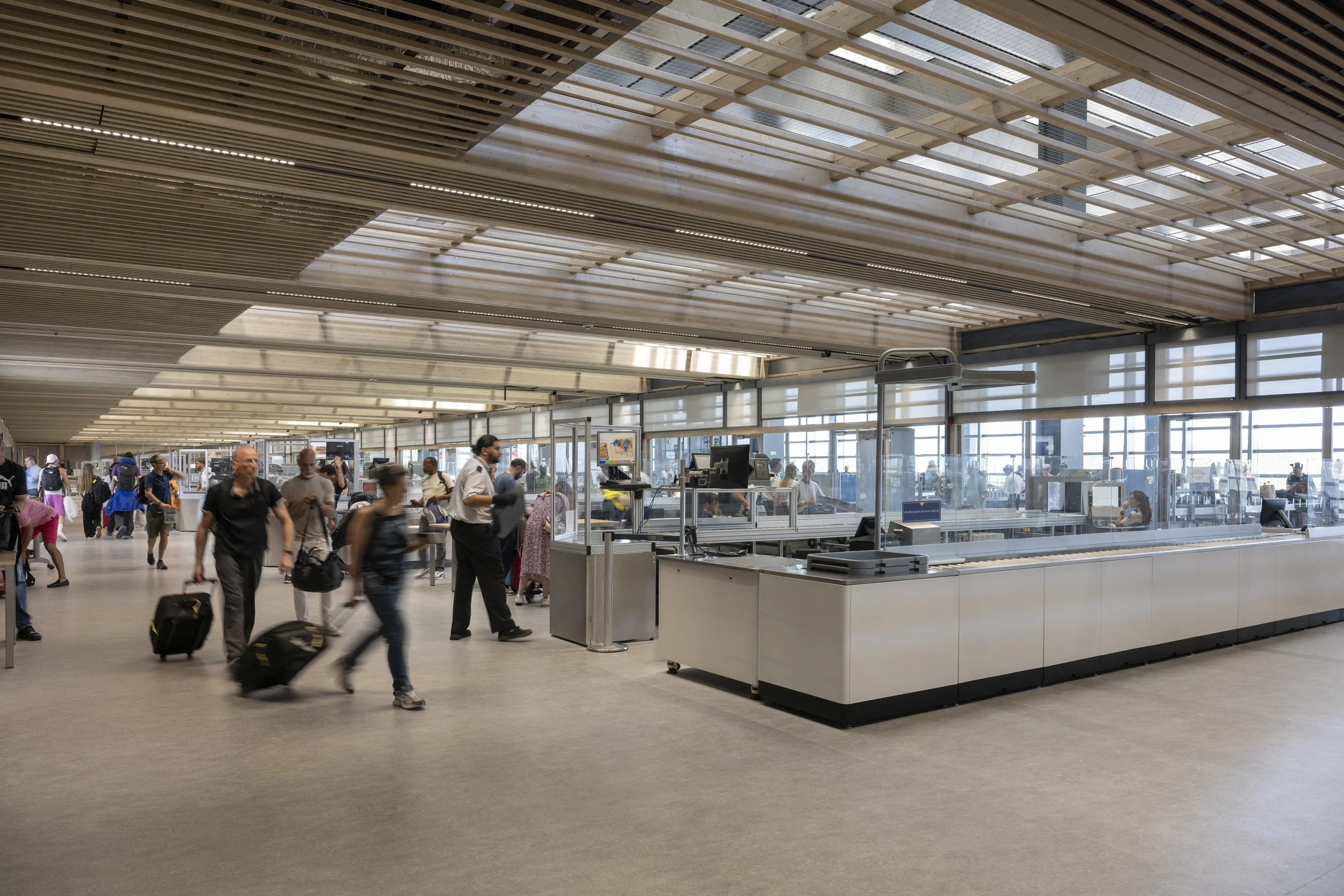 Passengers walking through an airport terminal with luggage, check-in counters, and large windows letting in natural light.