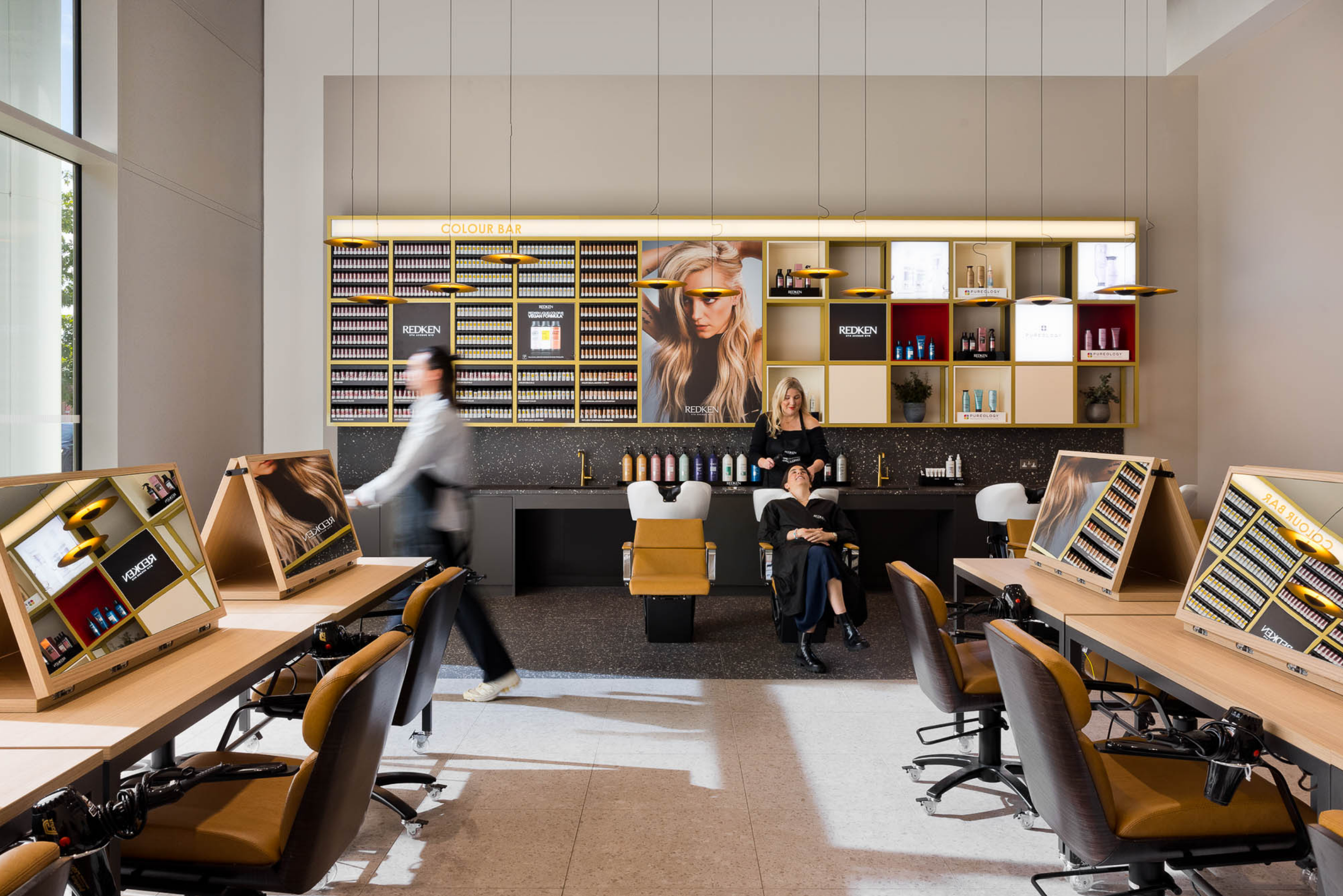 Interior of a modern hair salon with styling stations, a washing area with a stylist washing a woman's hair, and shelves stocked with hair products. Bright lighting and large windows provide natural light.
