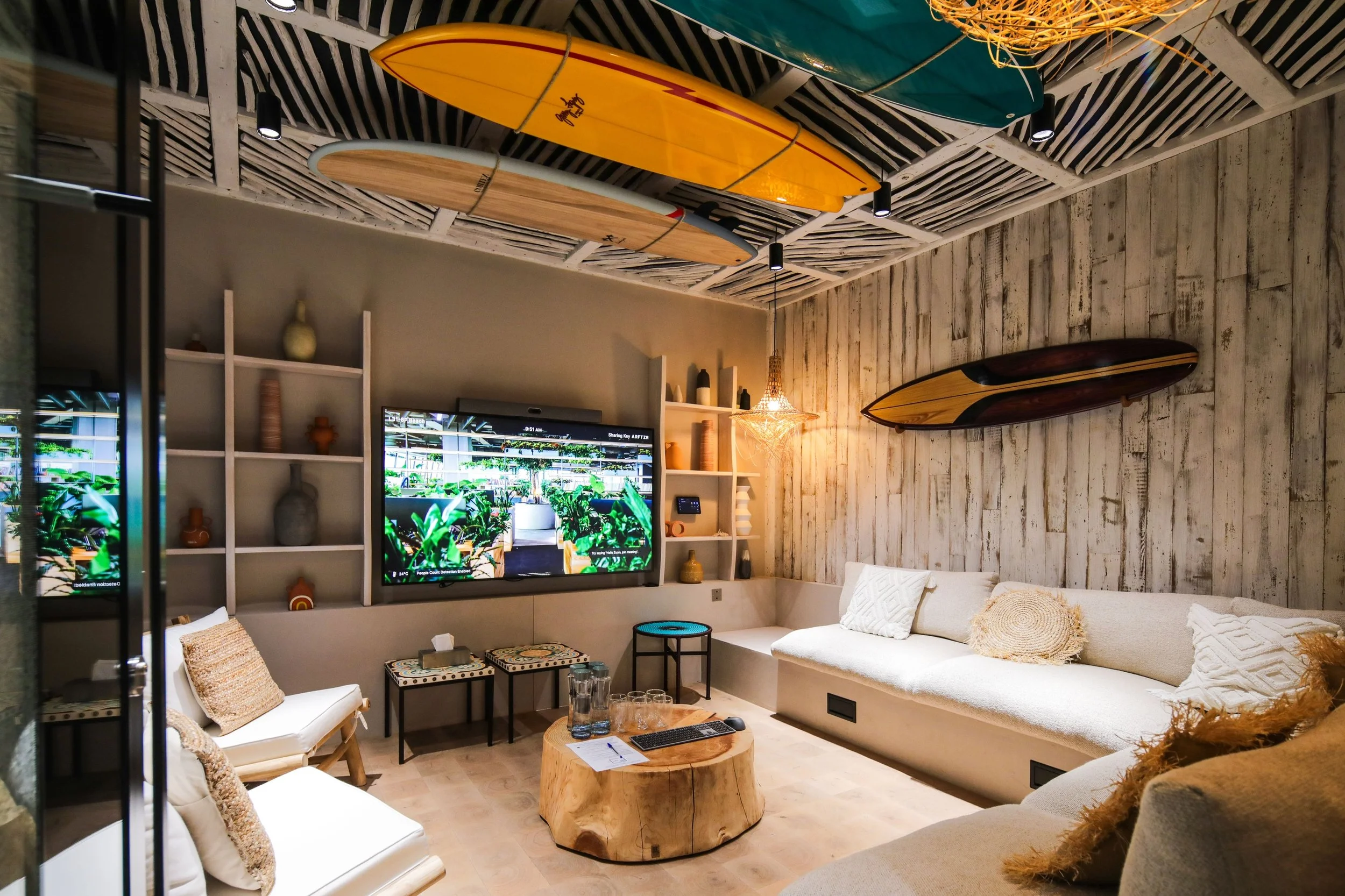 Living room with surfboards hanging from a wooden ceiling and wall, a mounted flat-screen TV, beige sofas with decorative pillows, a wooden coffee table with glasses and a remote, and a wall with rustic wood paneling.