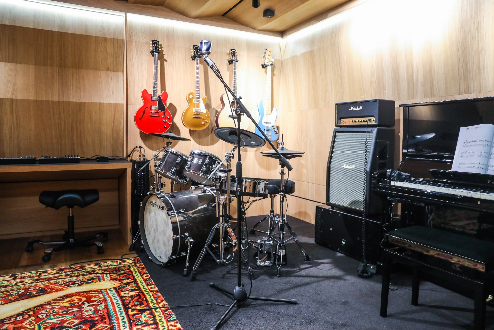 Music rehearsal space with guitars hanging on wooden wall, drum set in center, microphone and stand, keyboard on right, and recording equipment.