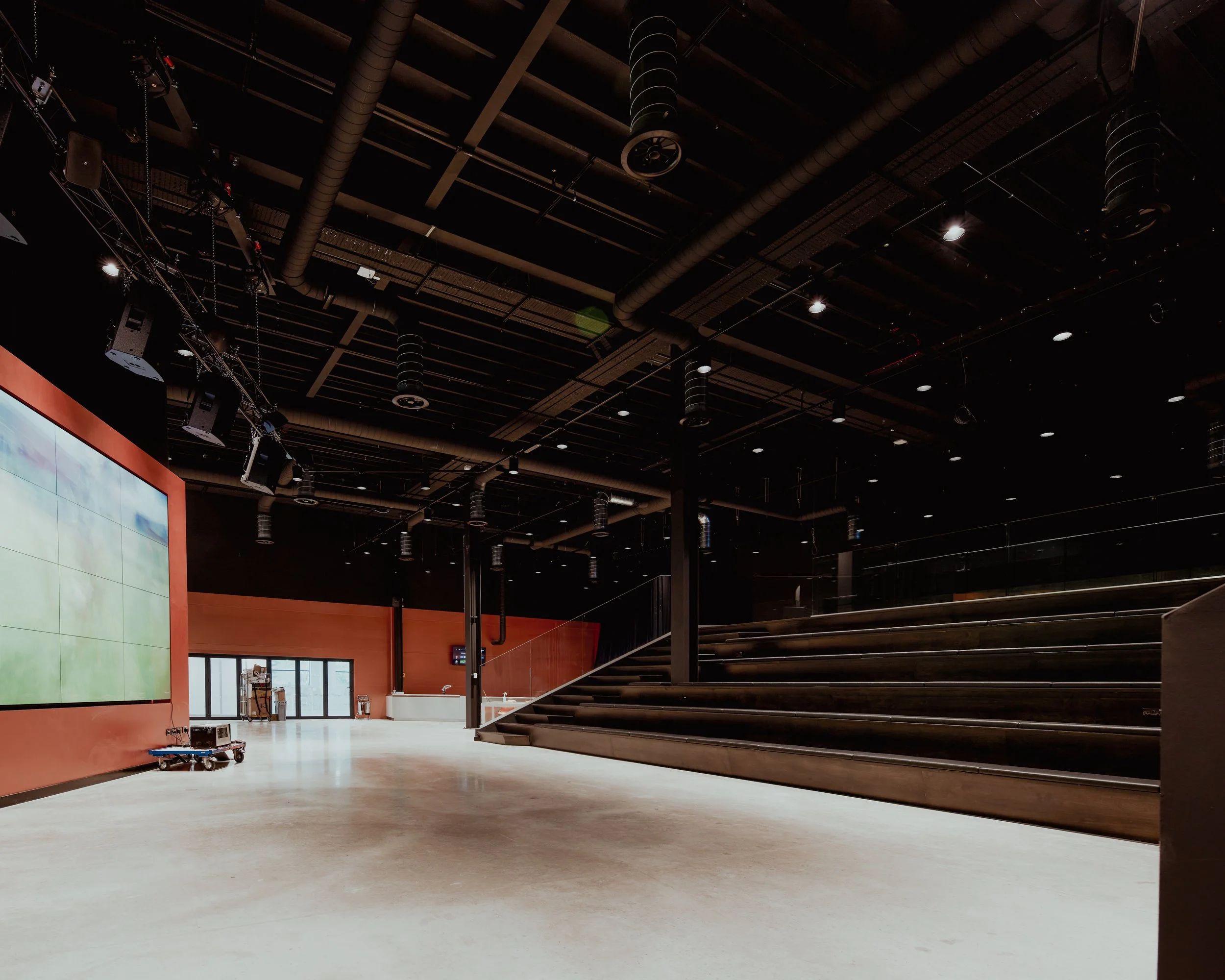 An empty, modern indoor auditorium or conference room with a large digital screen on the left wall, stairs on the right side, and a few doors and equipment in the background.