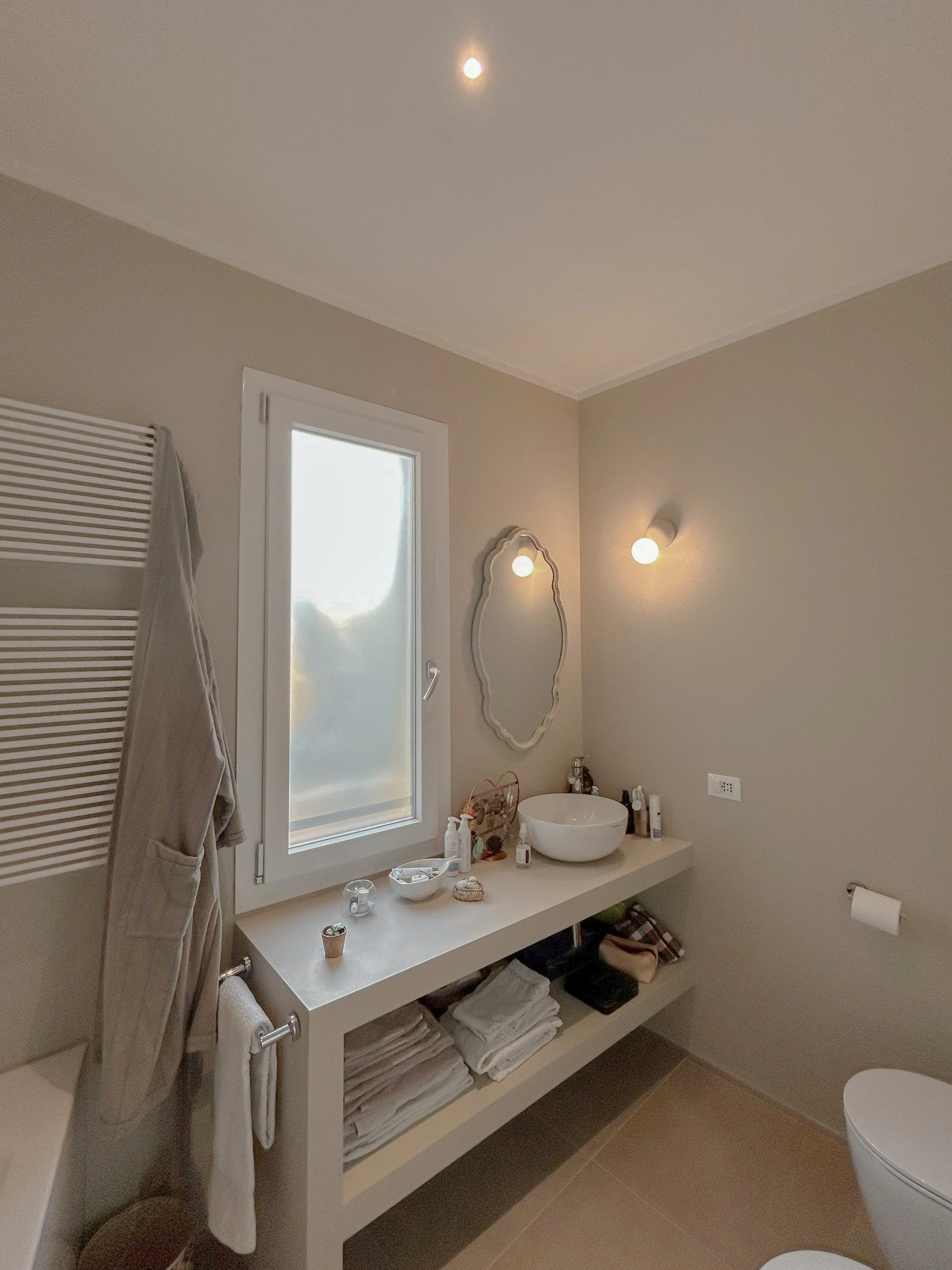 Modern bathroom with beige walls and a floating vanity with towels, toiletries, and a sink. There is a window, a wall-mounted mirror, and wall lights, with a towel rack and a partial view of a toilet.