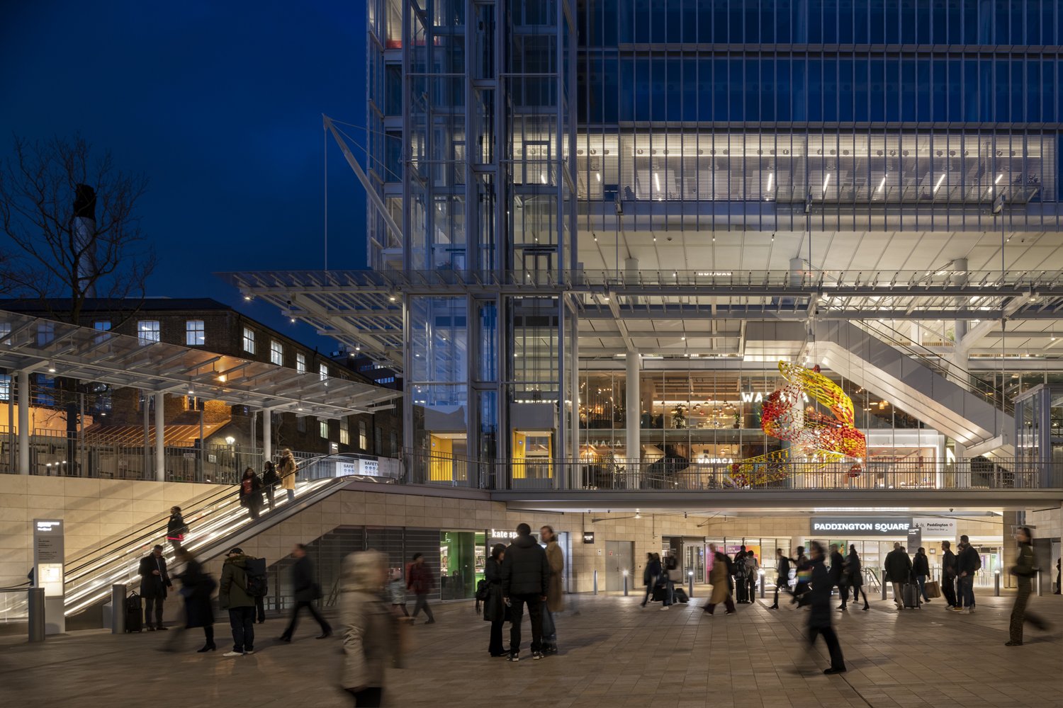 Night view of a busy train station, Paddington Square, with people walking and a modern glass building with colorful indoor decorations.