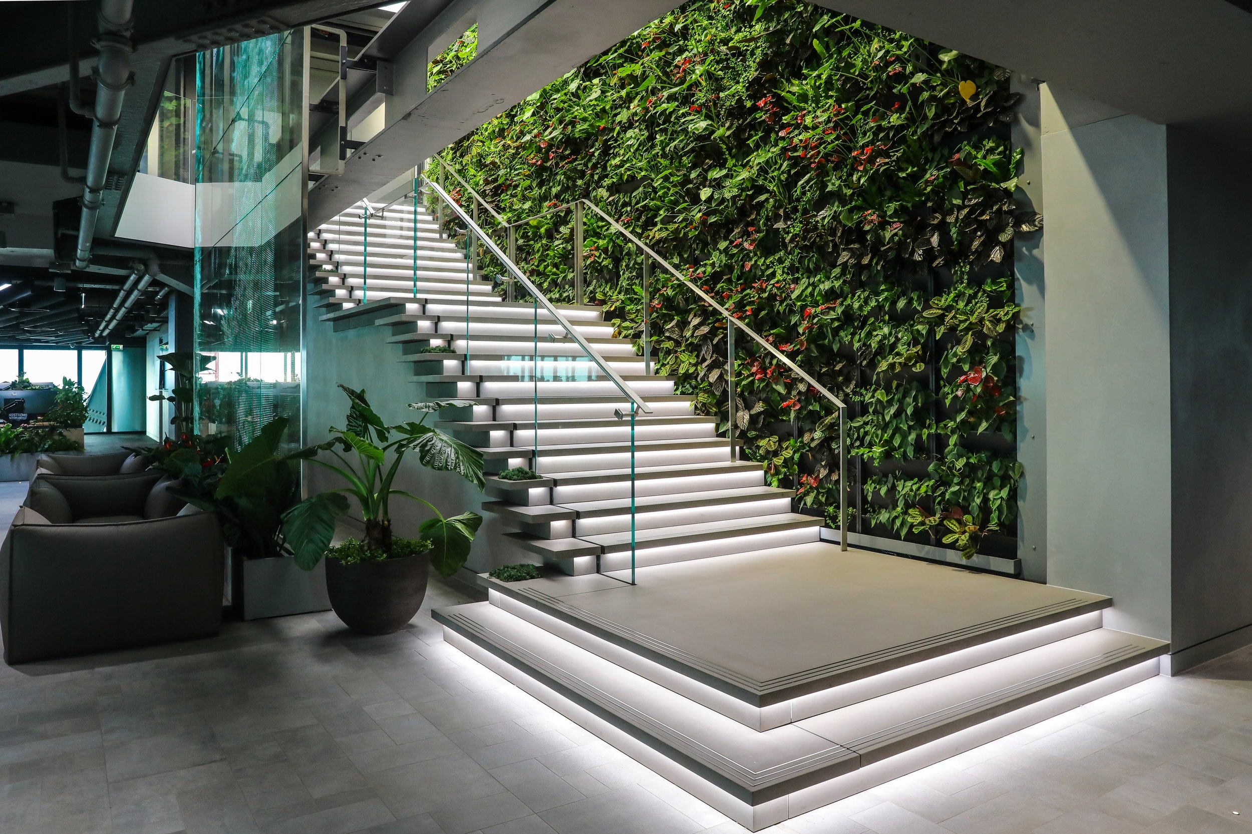 Modern indoor staircase with illuminated steps next to a vertical green wall filled with plants, in a contemporary office or lobby space.