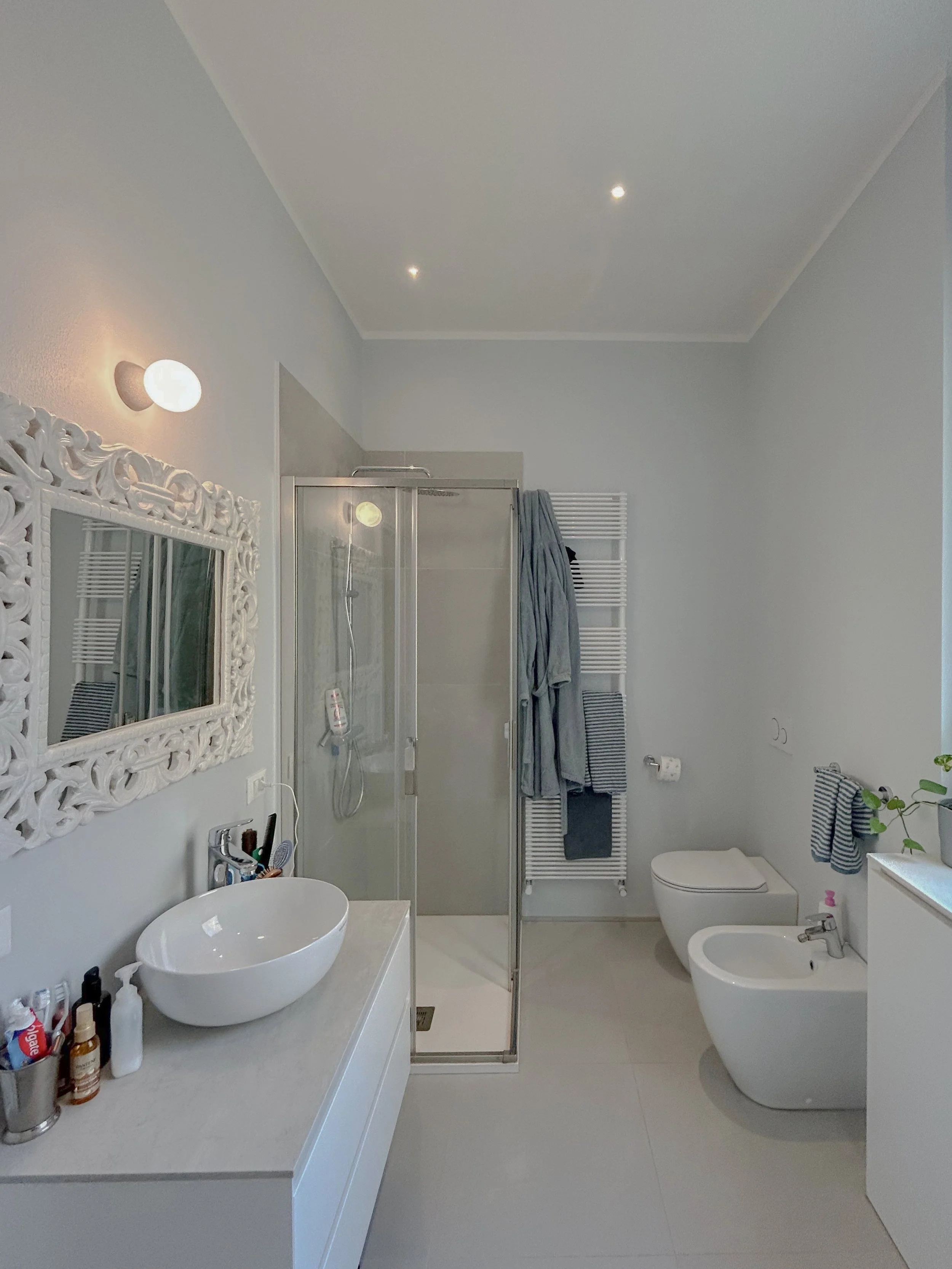 Modern bathroom with white walls, a decorative mirror, a vessel sink on a white vanity, a glass shower stall, a bidet, a toilet, and a heated towel rail with hanging towels.