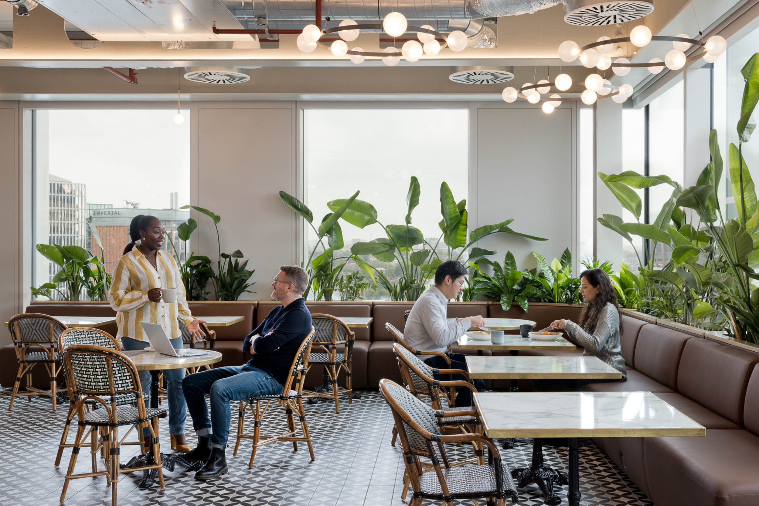 People sitting at tables and standing in a bright modern cafe with large windows, green plants, and patterned flooring, engaging in conversation and eating.