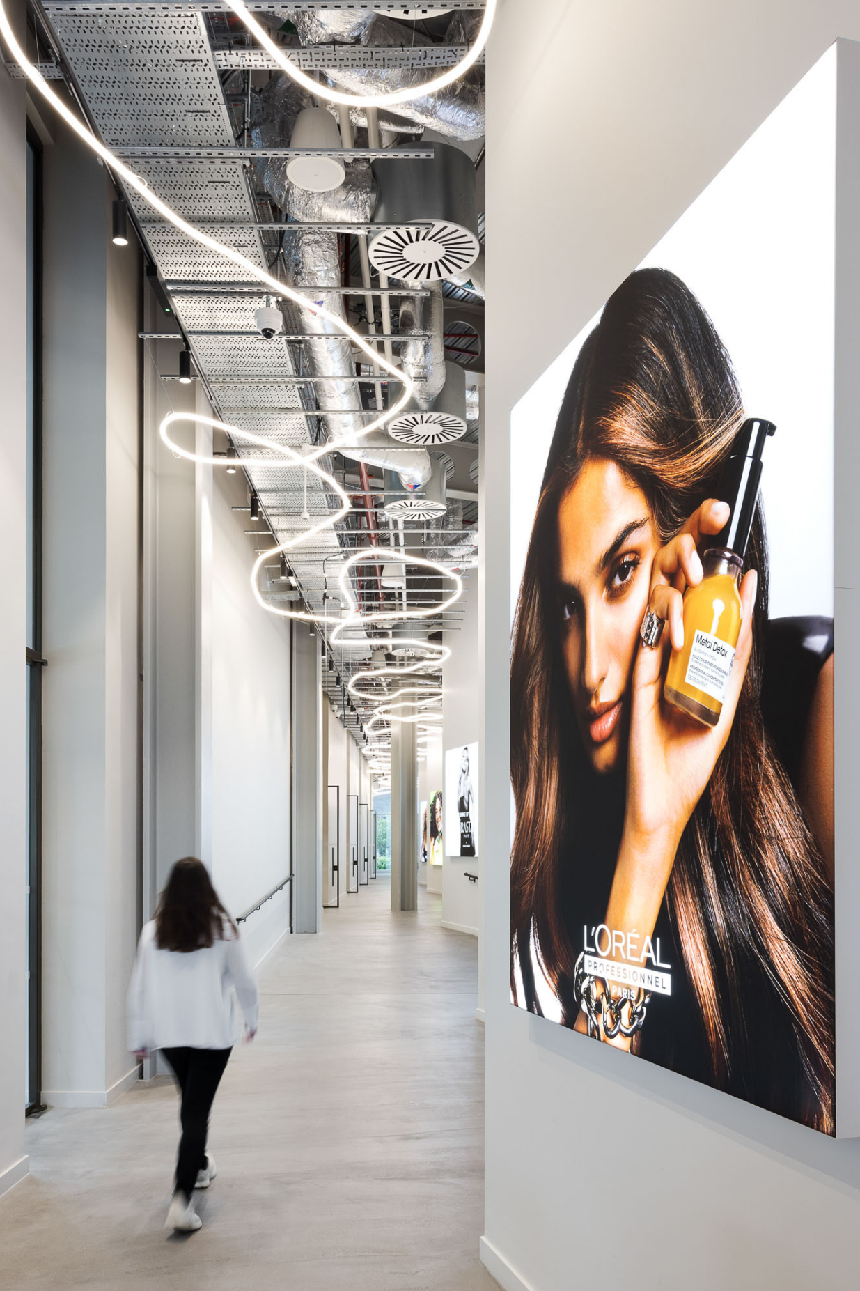 A hallway inside a modern building with large advertisement posters on the wall. One poster features a woman with long brown hair holding a hair product bottle. The ceiling has visible industrial ductwork and contemporary spiral lighting fixtures.