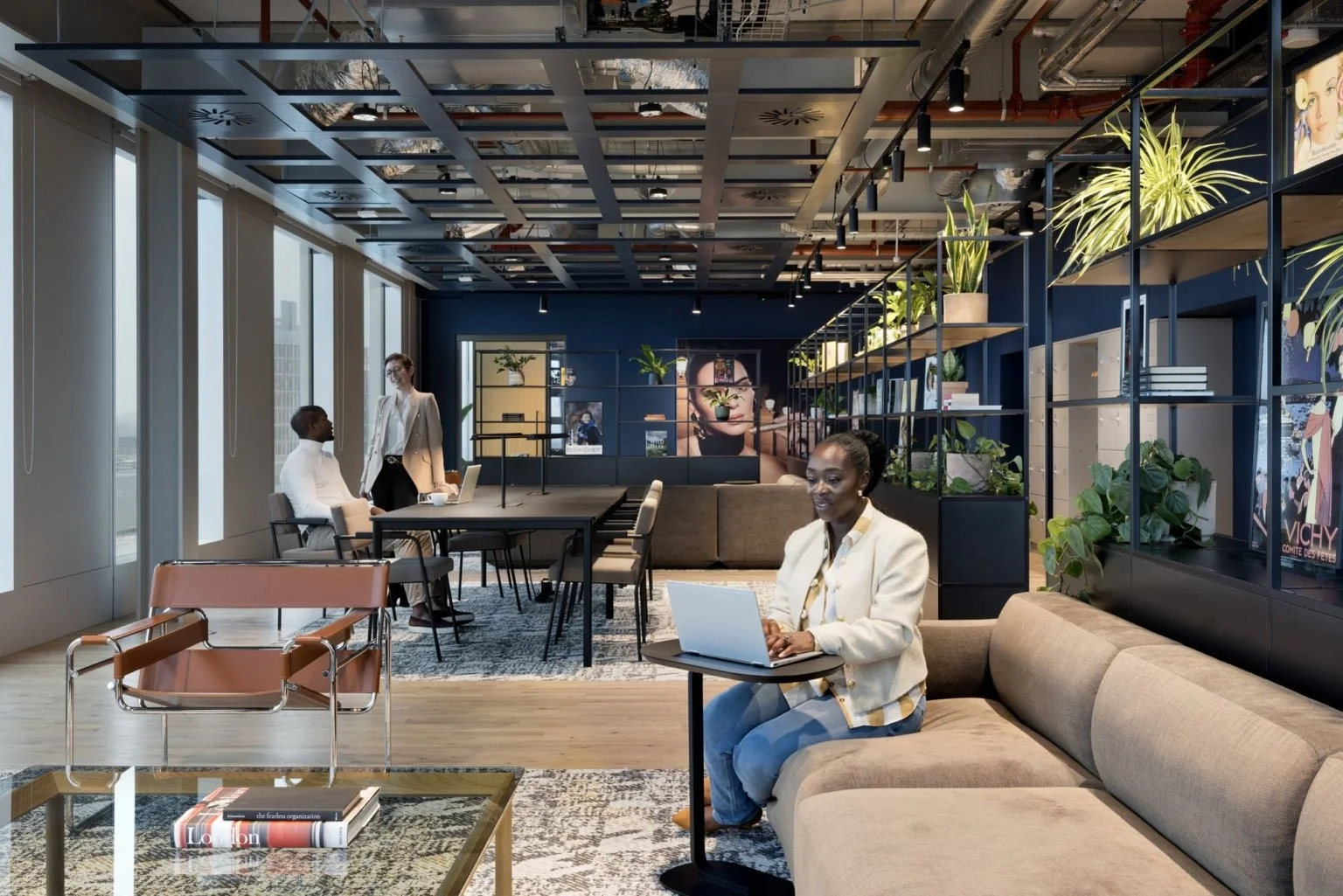 Modern office lounge with large windows, plants, and people working on laptops and having a discussion.