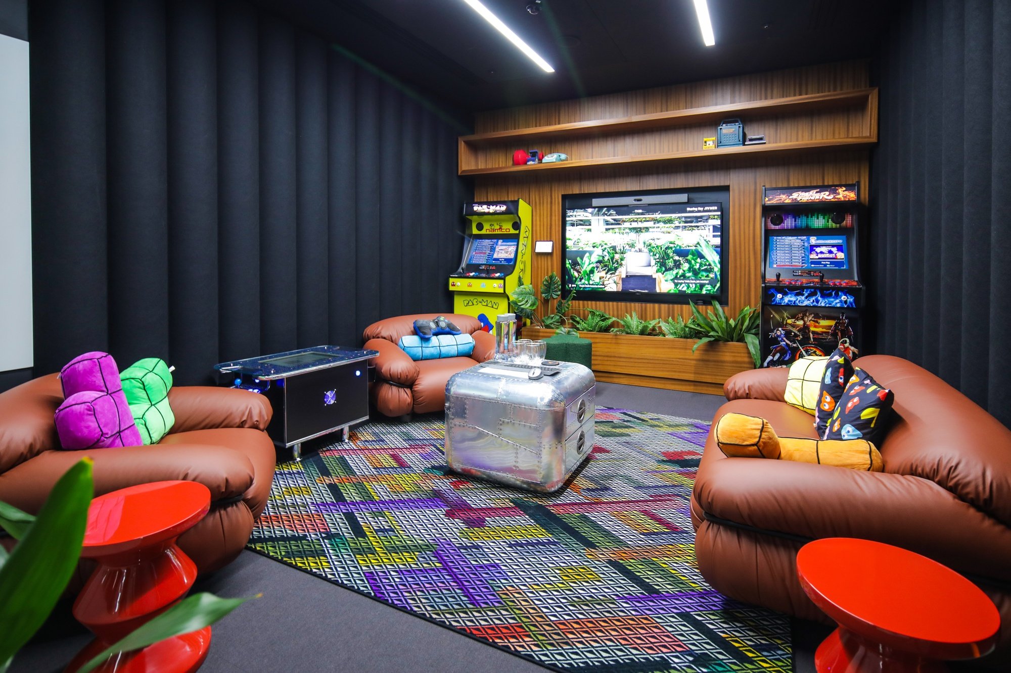 Colorful gaming room with three brown leather couches, arcade machines, a large TV, plants, and a vibrant patterned rug.