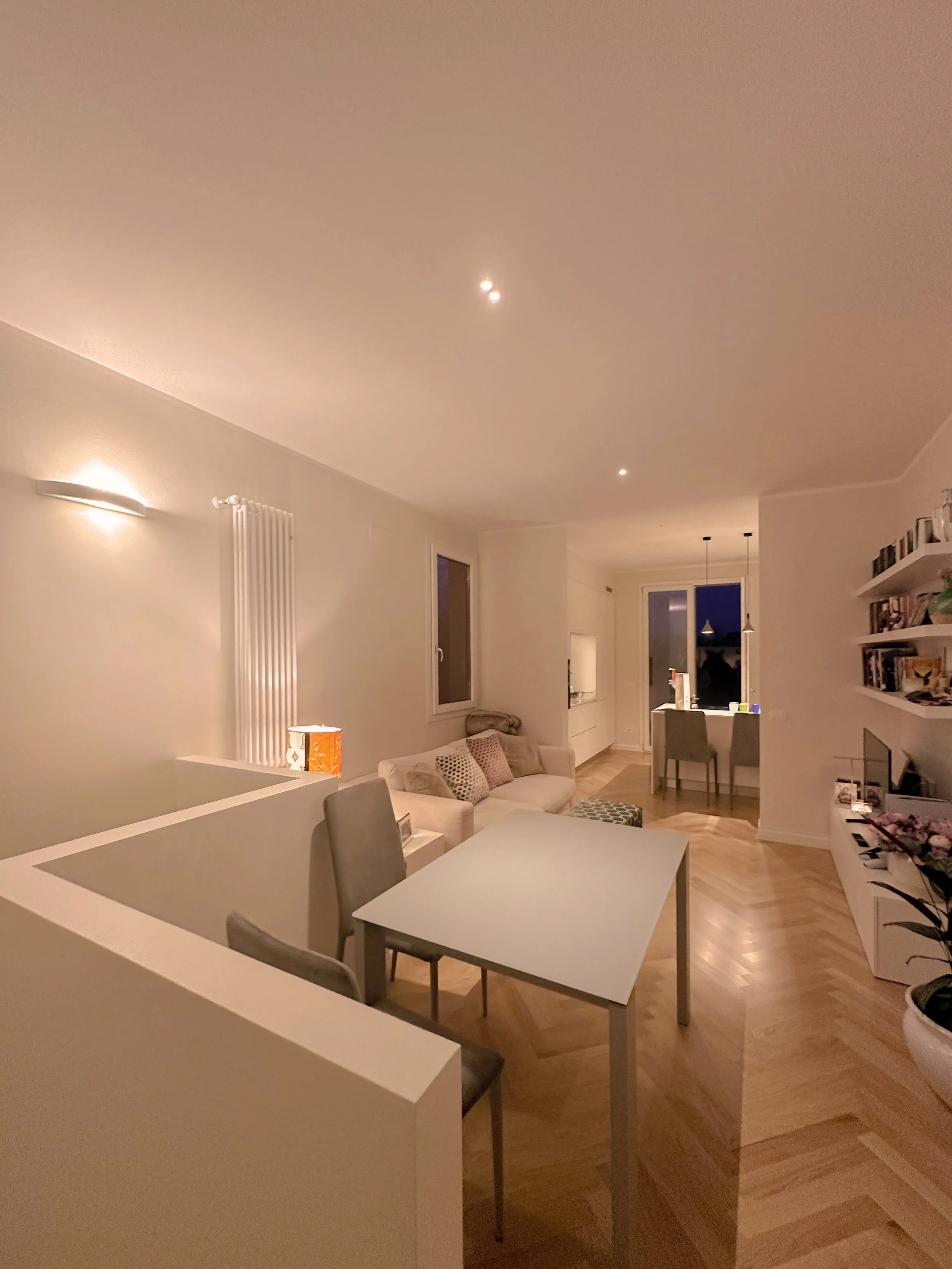 Living room with white walls, hardwood floors, a white couch with pillows, a table with books and a lamp, and large windows with evening sky visible.