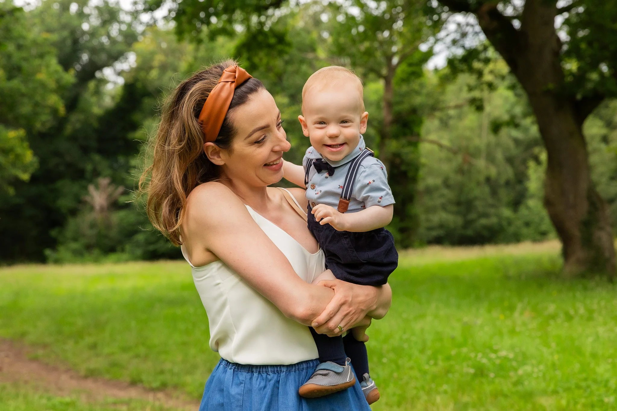A woman holding a baby boy outdoors in a grassy park with trees in the background.