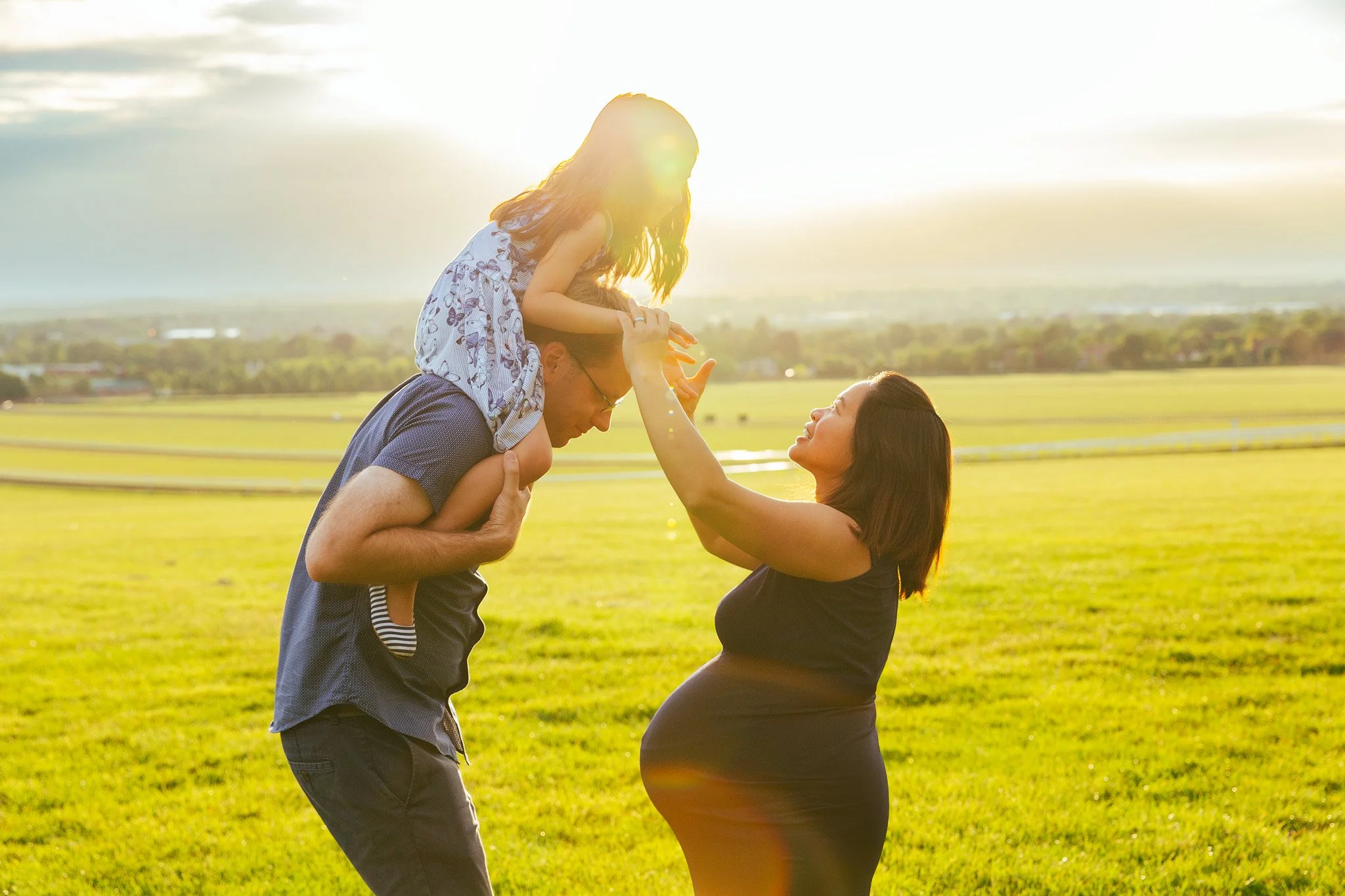 A family enjoying a moment outdoors at sunset. A pregnant woman stands facing a man carrying a young girl on his shoulders, who is reaching out to her. They are in a grassy field with a scenic horizon in the background.