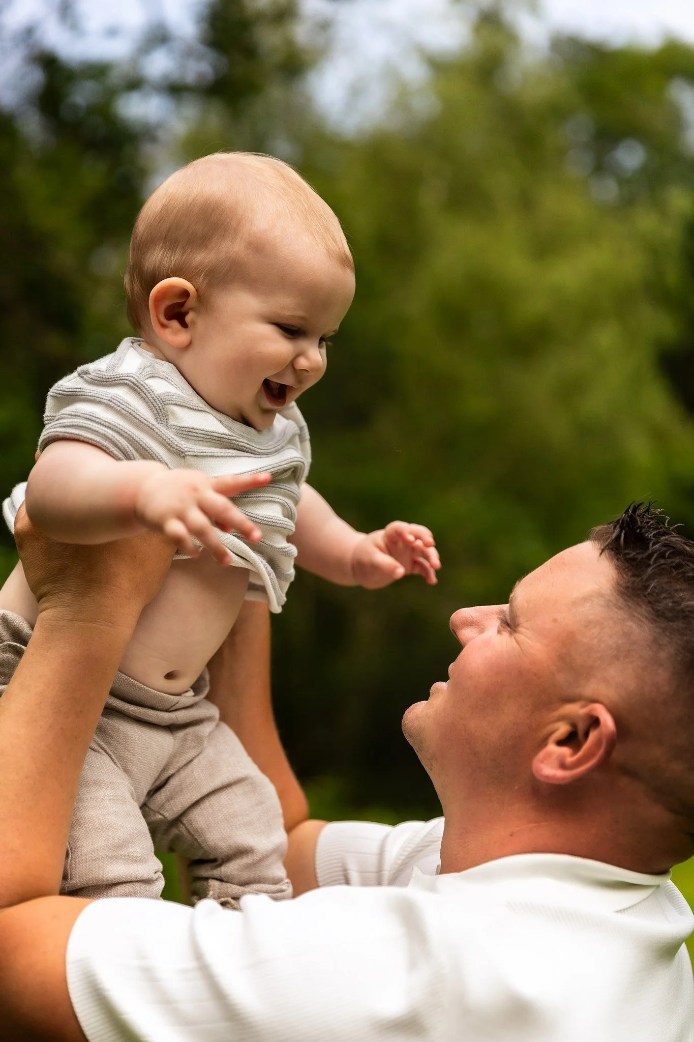 A man lifting a smiling baby in an outdoor park with green trees in the background.