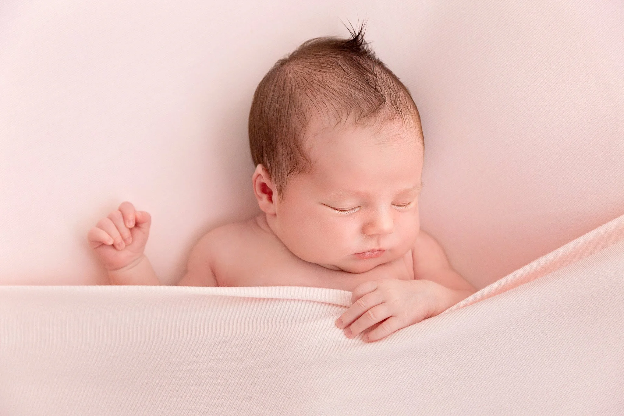 A peacefully sleeping newborn baby with closed eyes, light skin, and brown hair, lying on a soft pink surface with a matching pink blanket partially covering the body.