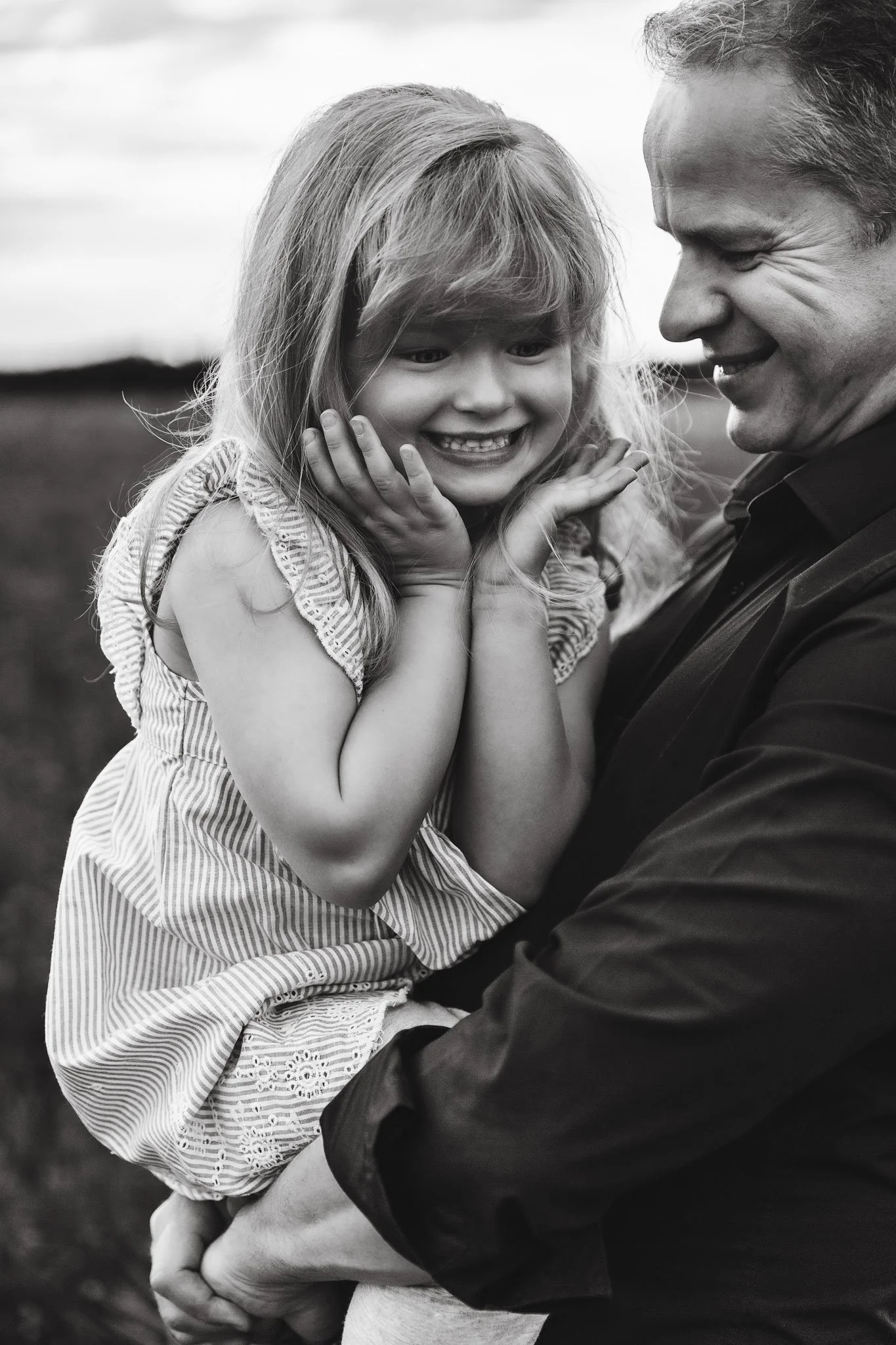 A man holding a young girl, both smiling and appearing joyful, outdoors with a blurry landscape in the background, in black and white.