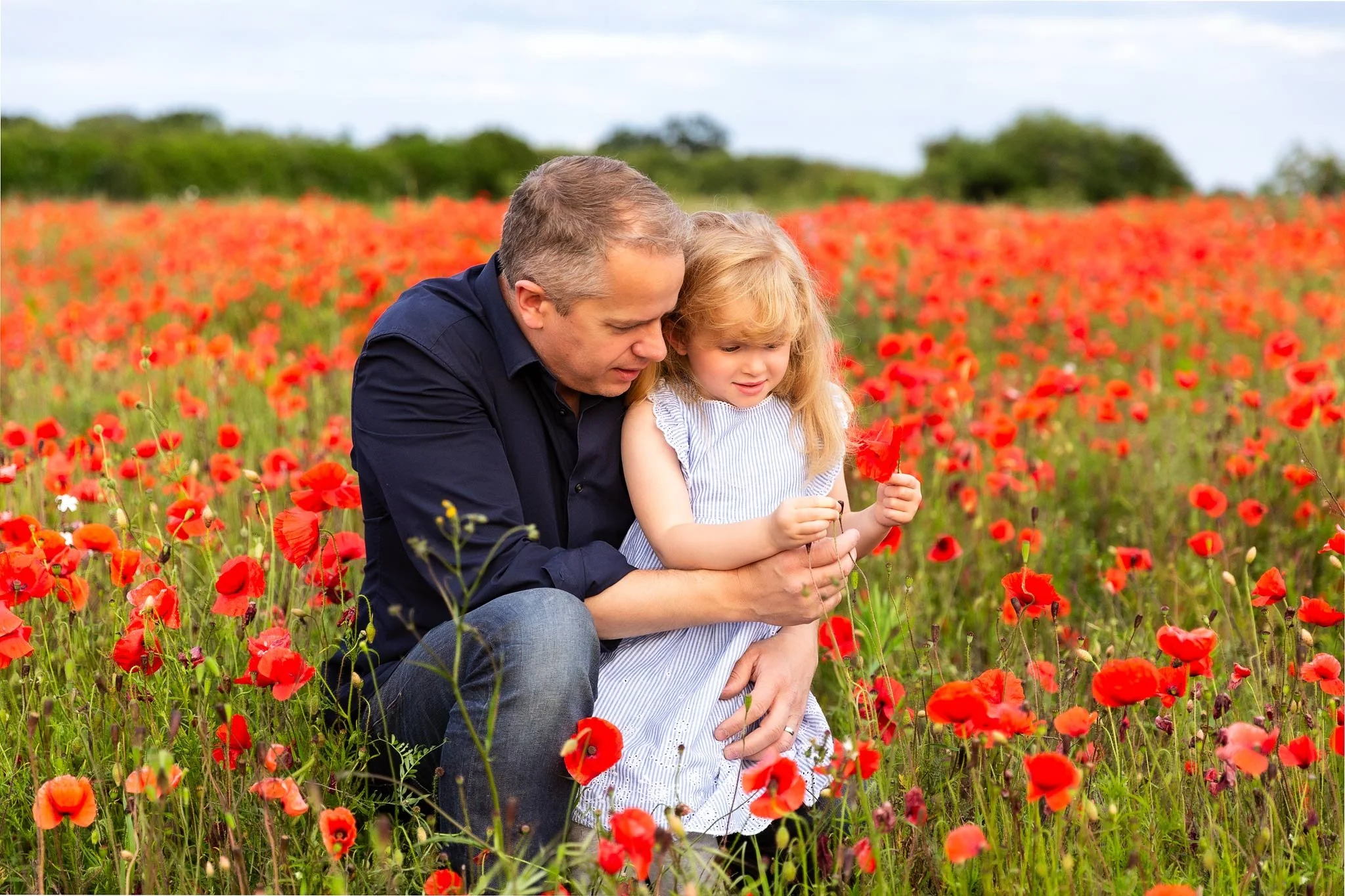 A man and a young girl in a field of red poppies, with the man kneeling and the girl holding a poppy and looking at it, during daytime.