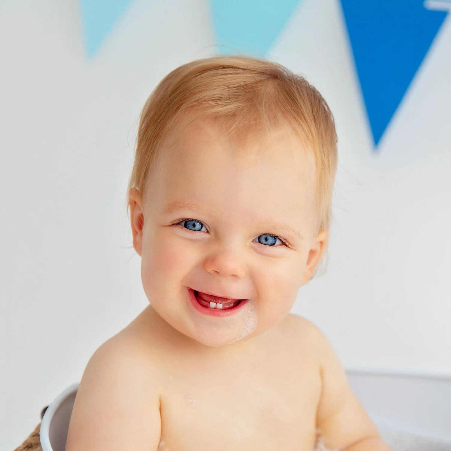 A cheerful little boy with short blond hair smiles at the camera, proudly showing his two front teeth.