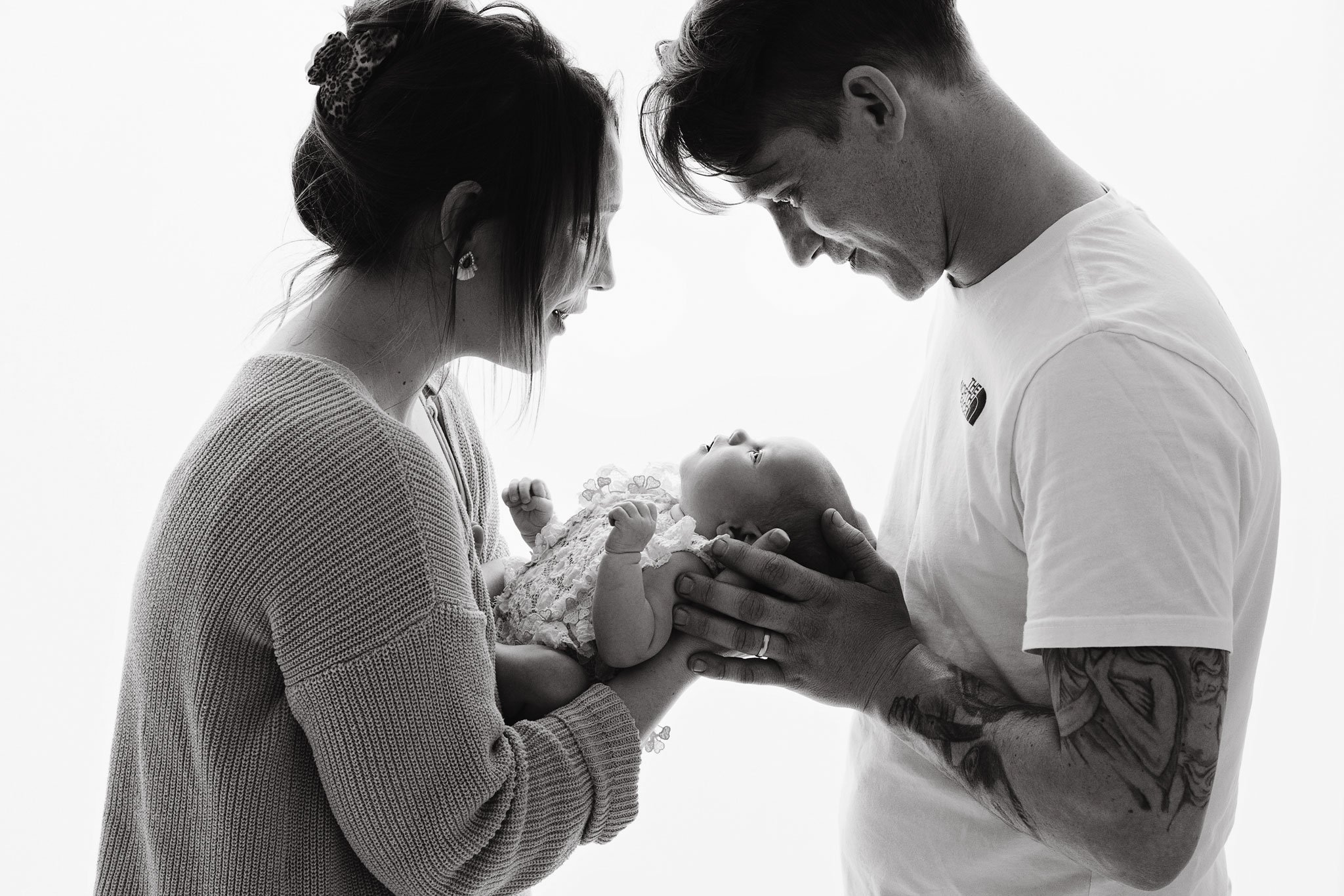 A couple holding an infant baby in a tender moment, with the woman on the left and the man on the right, both looking at the baby who is gazing up at them.