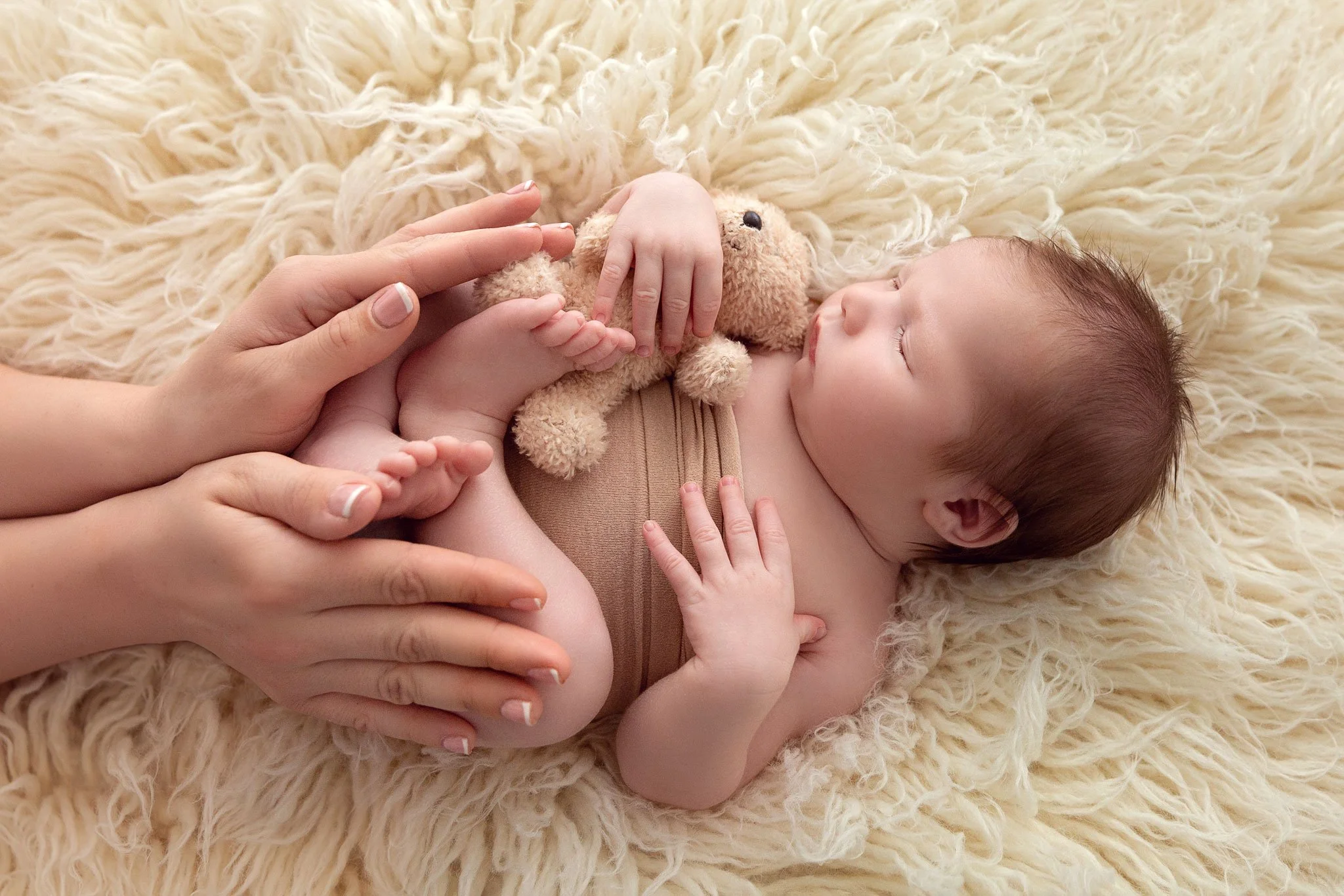 Baby lying on a plush beige rug holding a small teddy bear, with adult hands gently touching the baby’s feet.