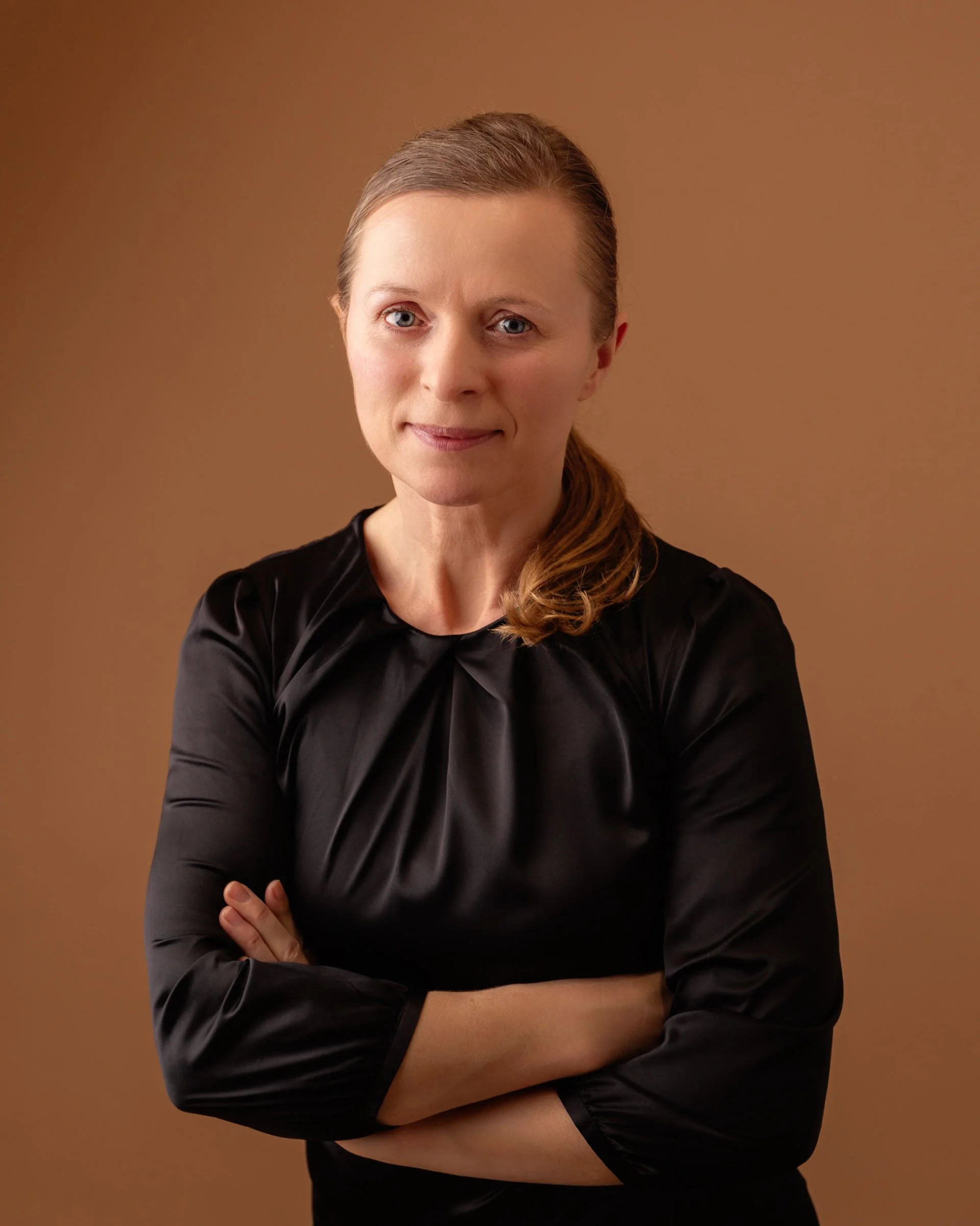 A woman with light skin, blue eyes, and light brown hair tied in a ponytail, wearing a black satin blouse with her arms crossed, standing against a plain brown background.