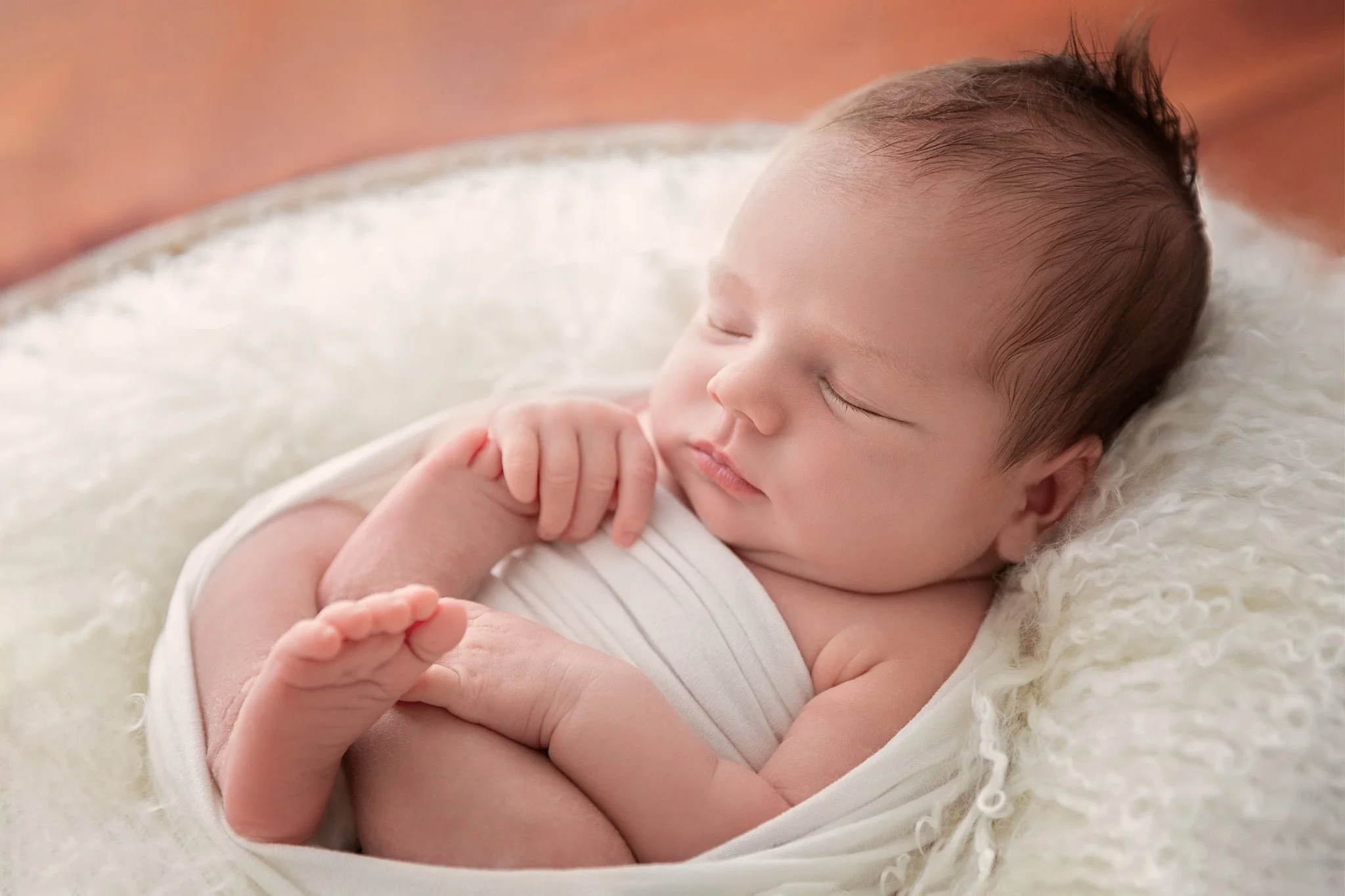 Close-up of a sleeping newborn baby wrapped in a white blanket, lying on a soft, fluffy surface.