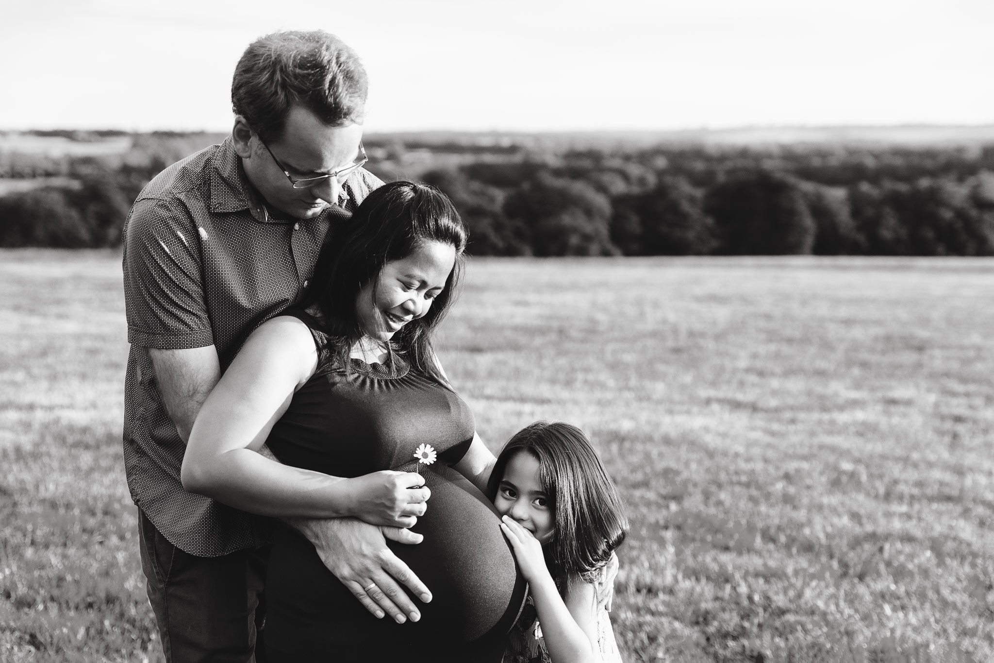 A black and white photo of a family outdoors in an open grassy field. A pregnant woman is holding her belly, with a young girl leaning on her and touching her belly. A man is standing behind them, resting his hands on the woman's belly, all smiling a