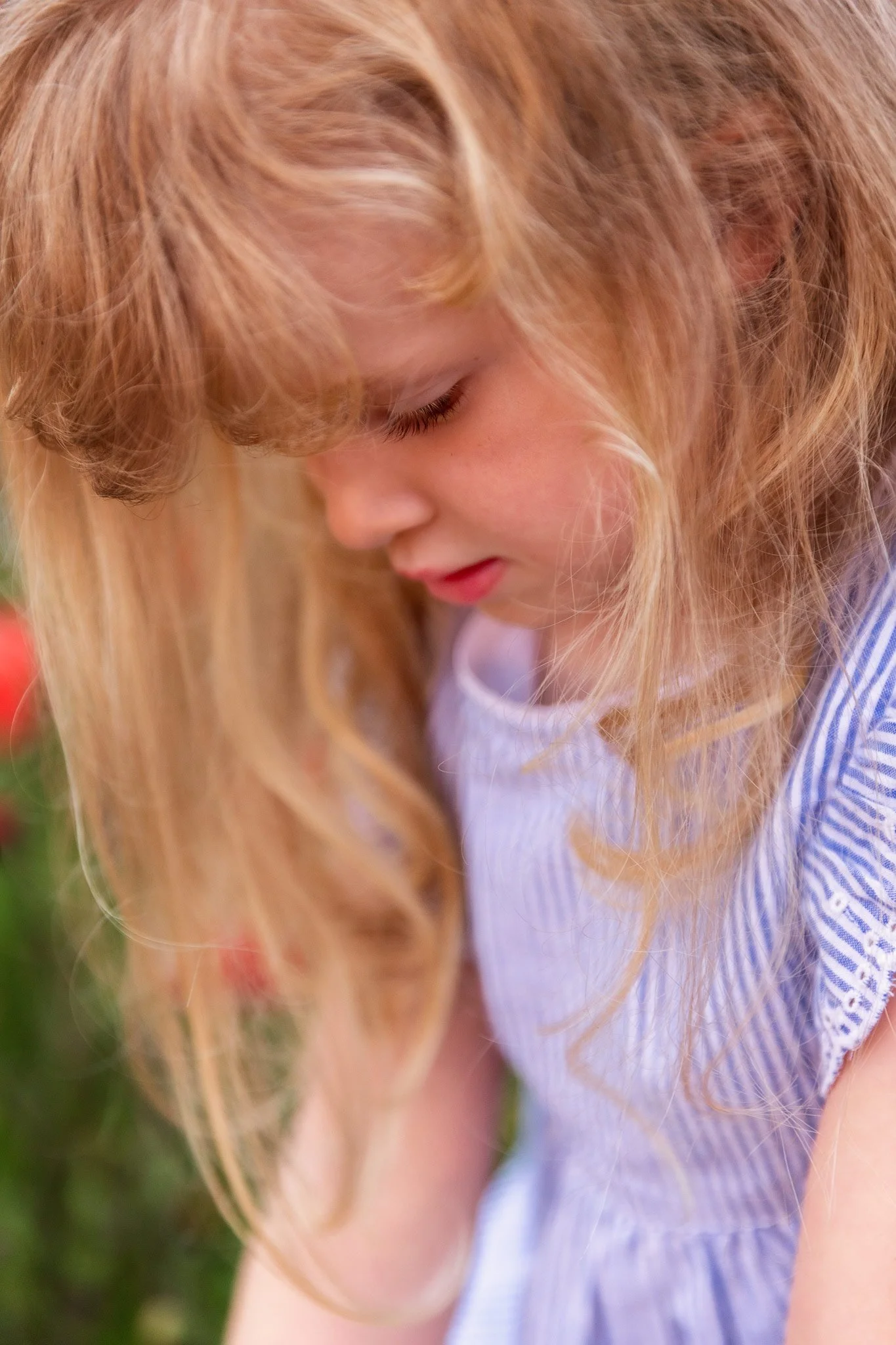 Close-up of a young girl with long, curly blond hair, looking downward, wearing a light blue striped dress, outdoors.